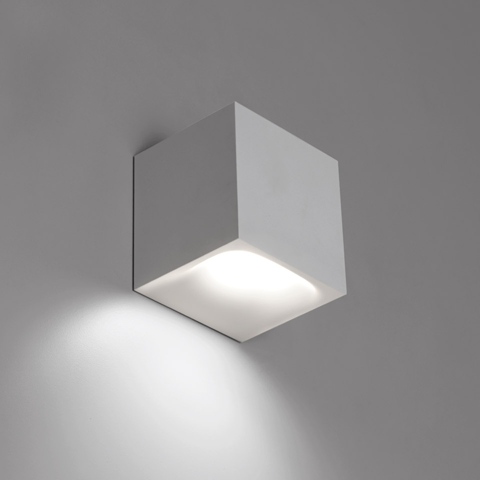 Artemide Aede wall light: compact white cubic LED sconce emitting warm downward light, mounted on grey wall