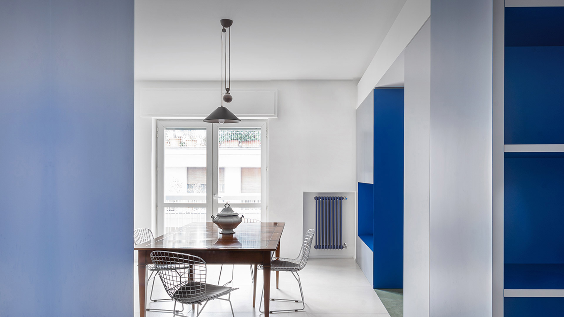 Dining room with Artemide Aggregato pendant lamp in dark metal over a wooden table with wire chairs, blue shelving unit on the right