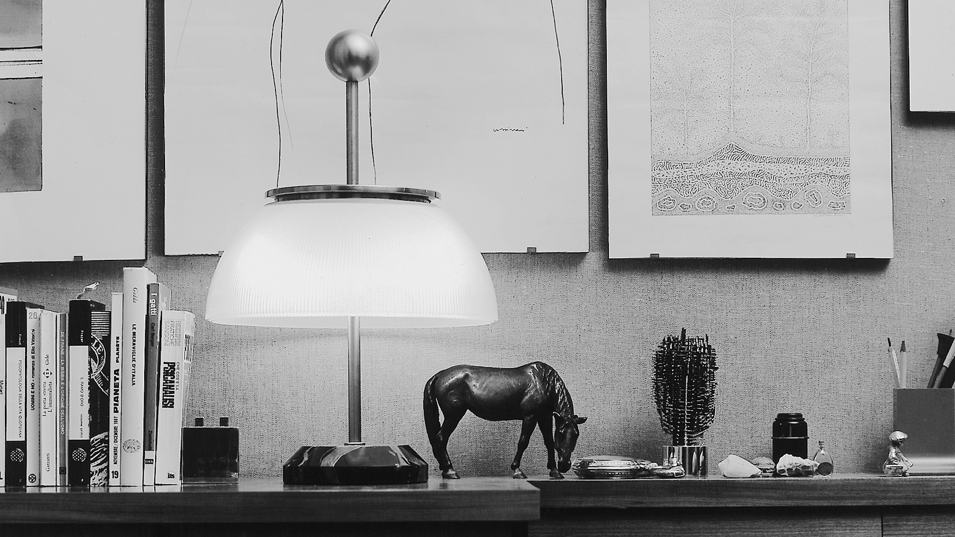 Lampada da tavolo Artemide Alfa con paralume a cupola in vetro opalino e struttura in metallo scuro, che diffonde luce calda su un comodino in legno