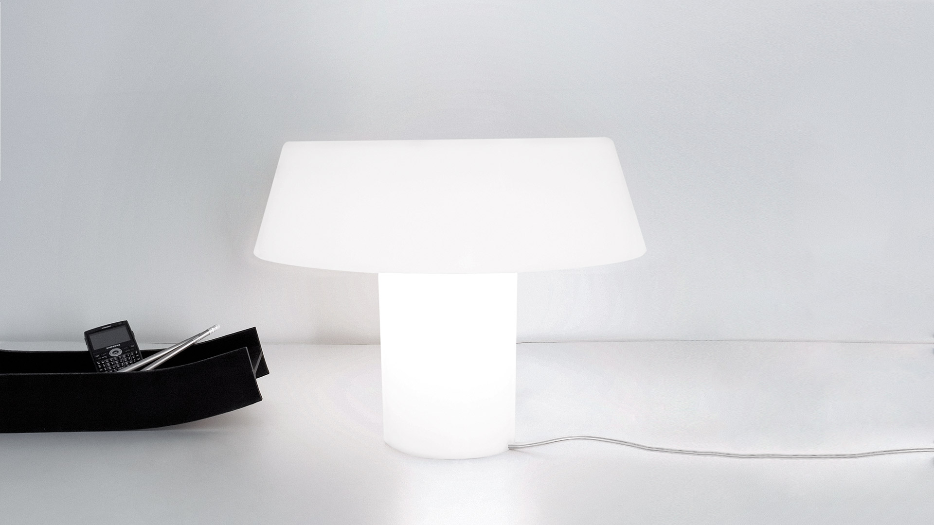 Artemide Amami table lamp with white opal cylindrical base and wide flat shade, emitting soft diffused light on a white surface