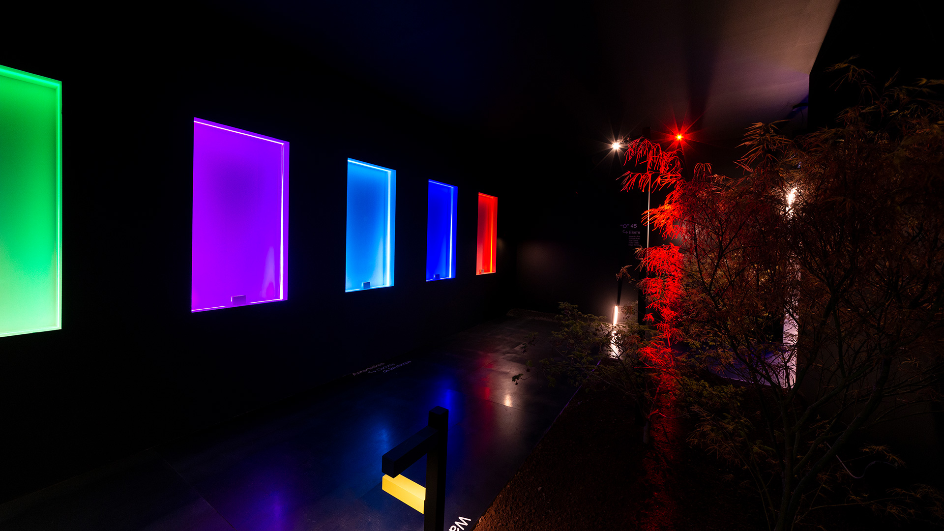Installazione esterna Artemide Antarktikós: pannelli rettangolari illuminati colorati in verde, viola, blu e rosso su sfondo scuro con fogliame illuminato