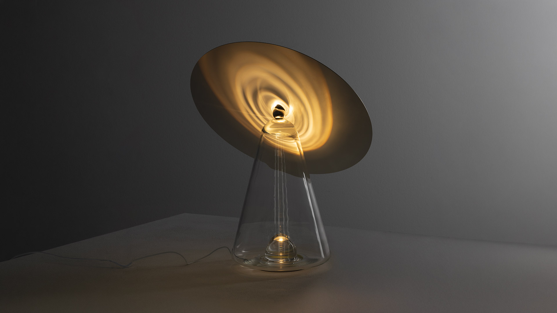 Lampada da tavolo Artemide Boltons: riflettore a disco dorato inclinato su base conica in vetro trasparente con sorgente luminosa calda