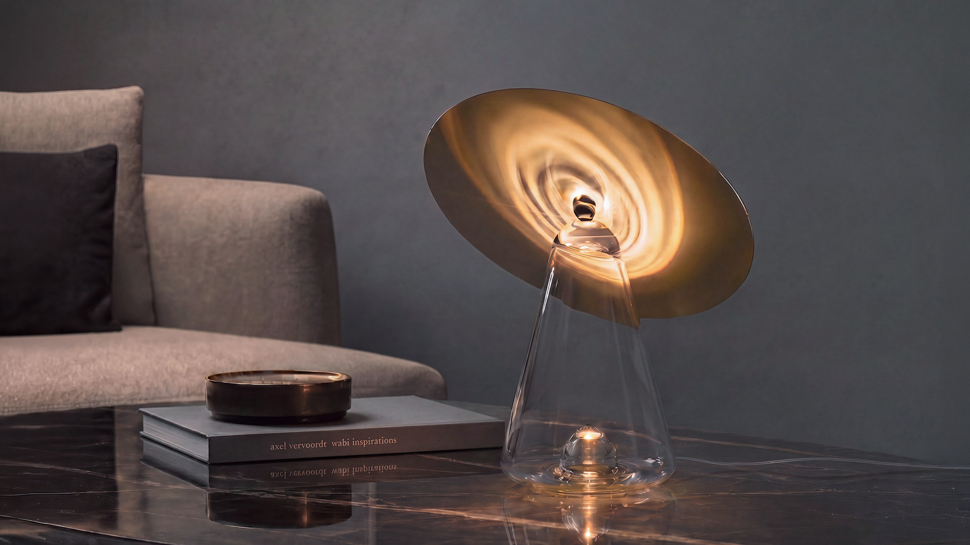Lampada da tavolo Artemide con base conica in vetro trasparente e paralume a disco bianco inclinato, accesa su un tavolino in marmo in un soggiorno moderno.