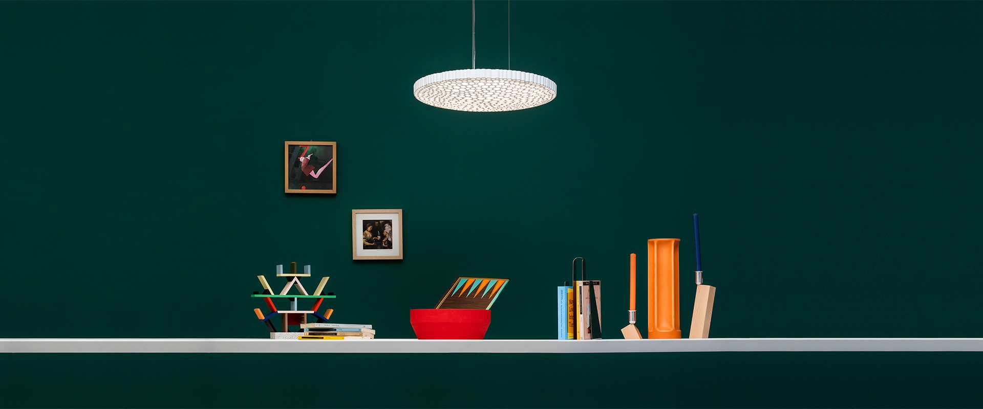 Lampada a sospensione Artemide Calipso con paralume a disco bianco perforato sopra uno scaffale con libri, oggetti colorati e quadri su parete verde scuro