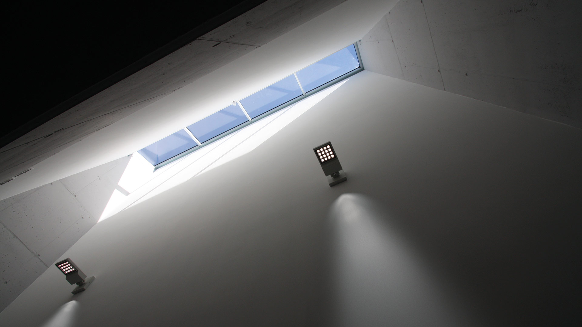 Due faretti LED a parete Artemide Cefiso illuminano una parete angolare bianca sotto un lucernario stretto con luce naturale.