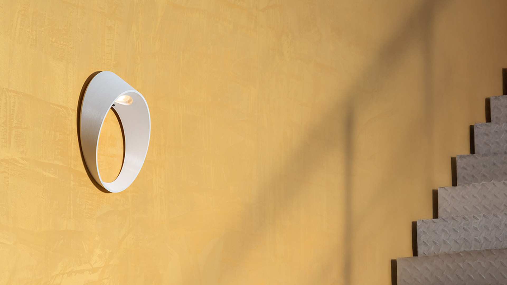 Lampada da parete Artemide Cleomede, diffusore circolare ad anello color tortora che emette luce calda, montata su parete scura con pianta