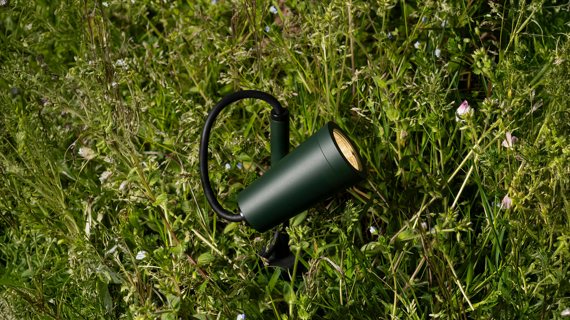 Faretto da esterno Artemide Conus in verde scuro, montato su picchetto in un prato rigoglioso, con luce calda orientata verso l'alto