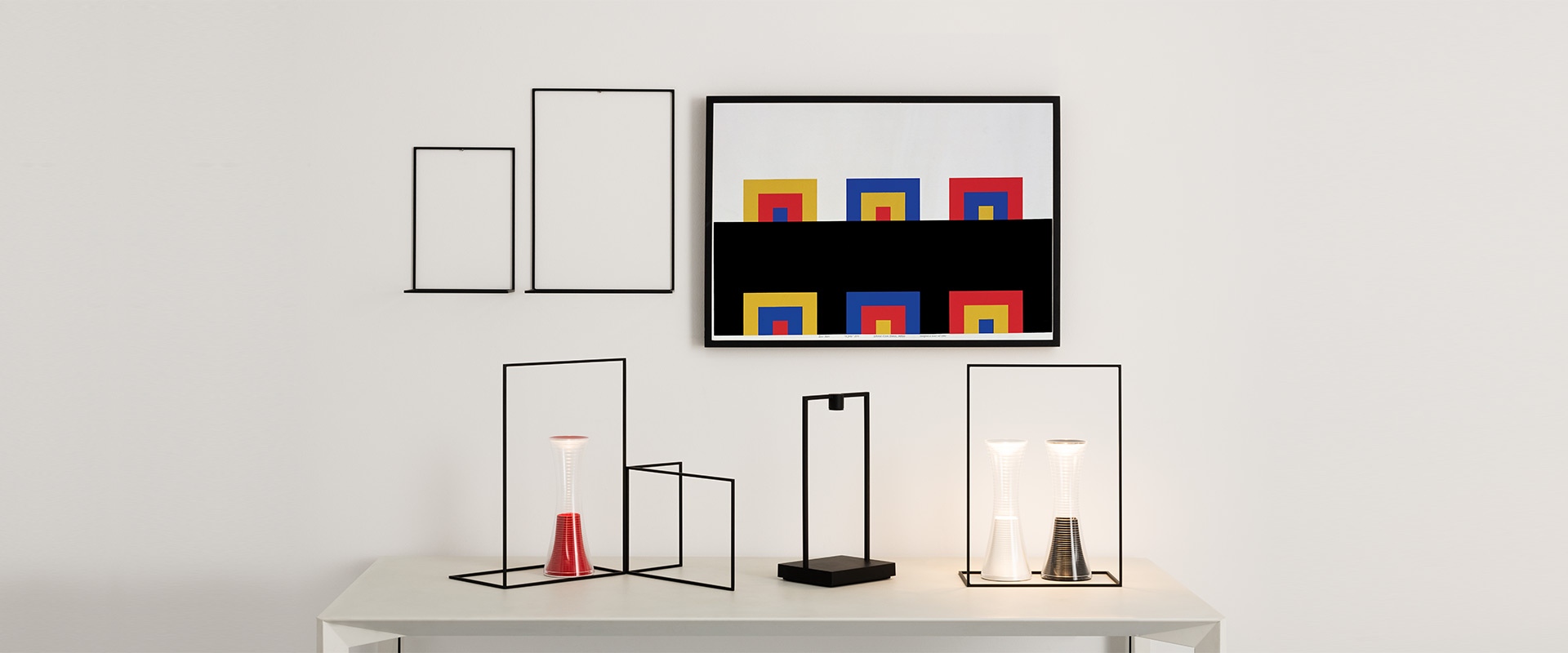 Lampada Artemide Curiosity: struttura rettangolare nera con luce LED integrata alla base, illumina una pianta, ciotole in ceramica e una brocca di vetro su superficie bianca