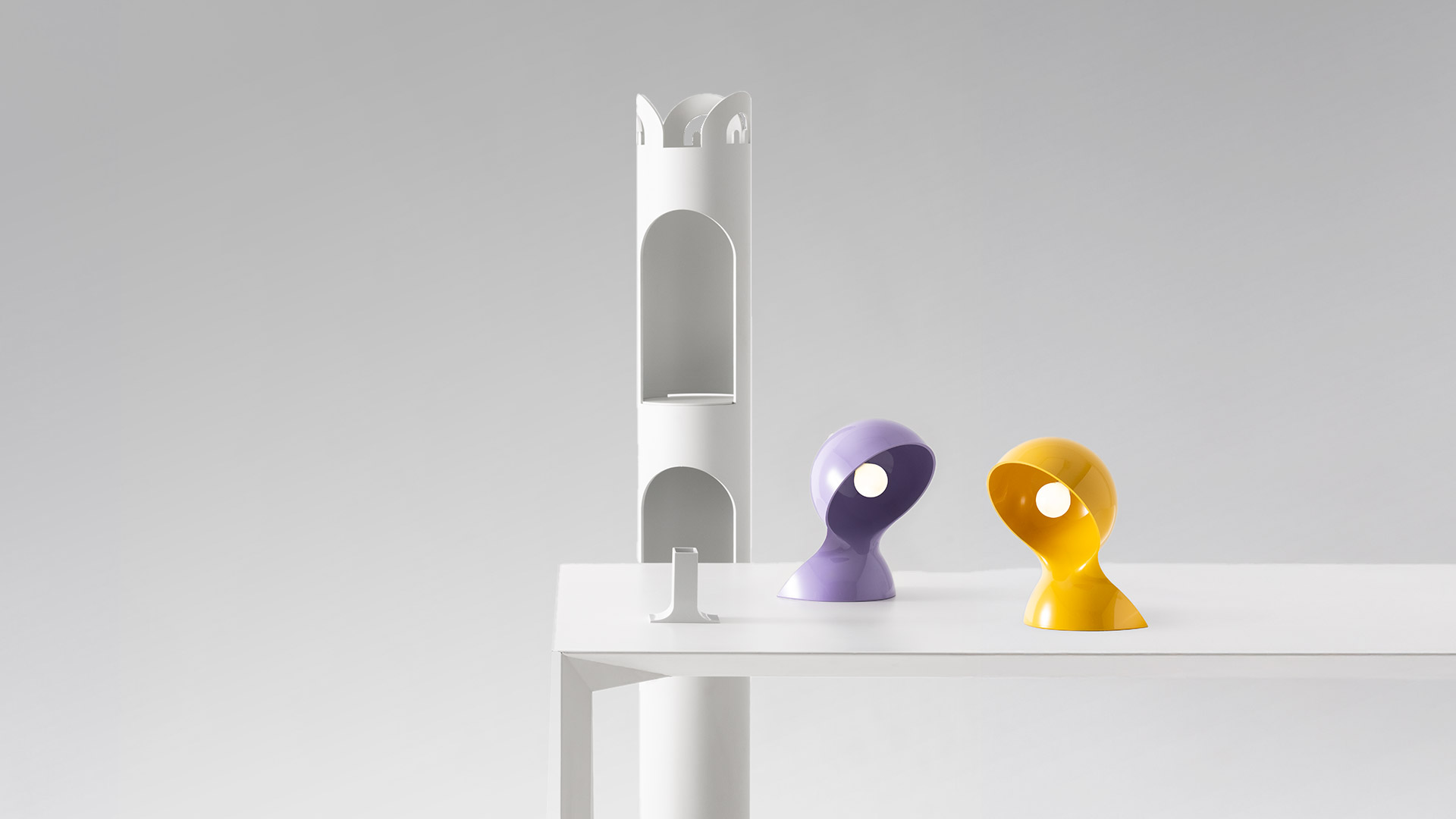 Lampada da tavolo Artemide Dalù bianca con forma organica curva, illuminata calorosamente su libri impilati accanto a fiori bianchi e una pianta
