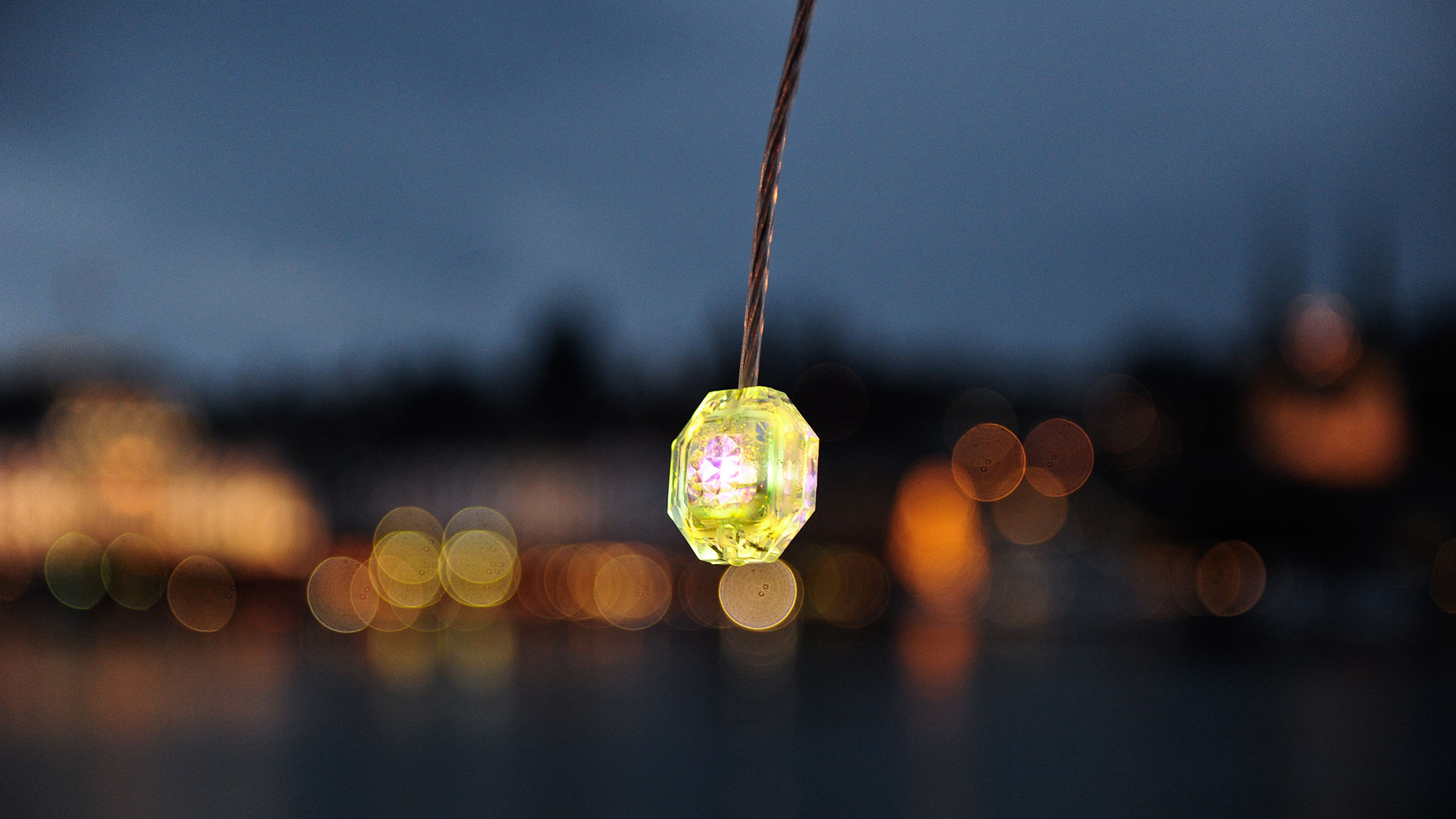 Lampada a sospensione Diamante di Artemide: piccola luce LED a forma di gemma sfaccettata giallo-verde appesa su paesaggio urbano al crepuscolo sfocato