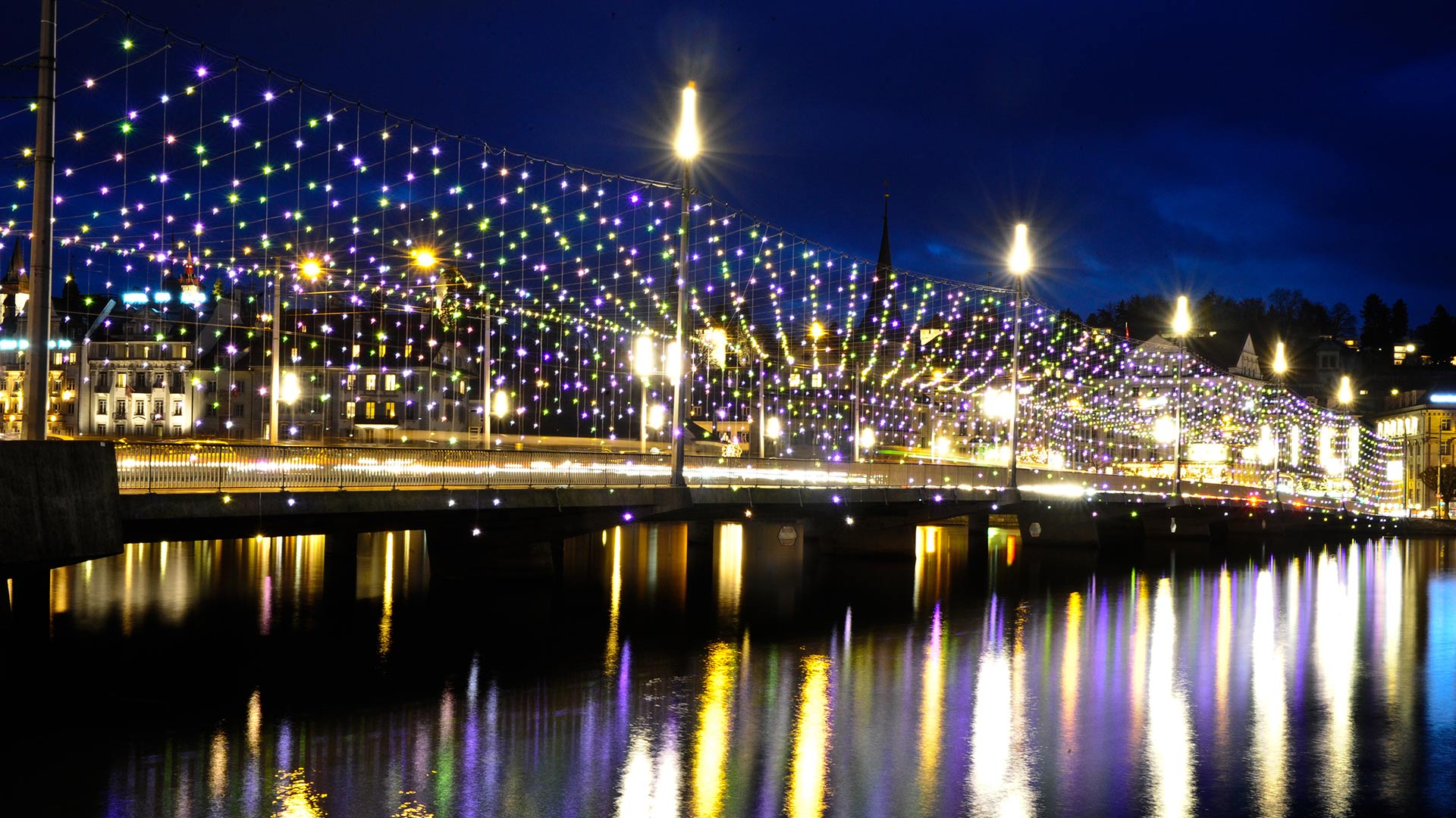 Ponte illuminato di notte con luci a stringa multicolori Artemide Diamante riflesse nel fiume sottostante, creando un'atmosfera urbana festiva