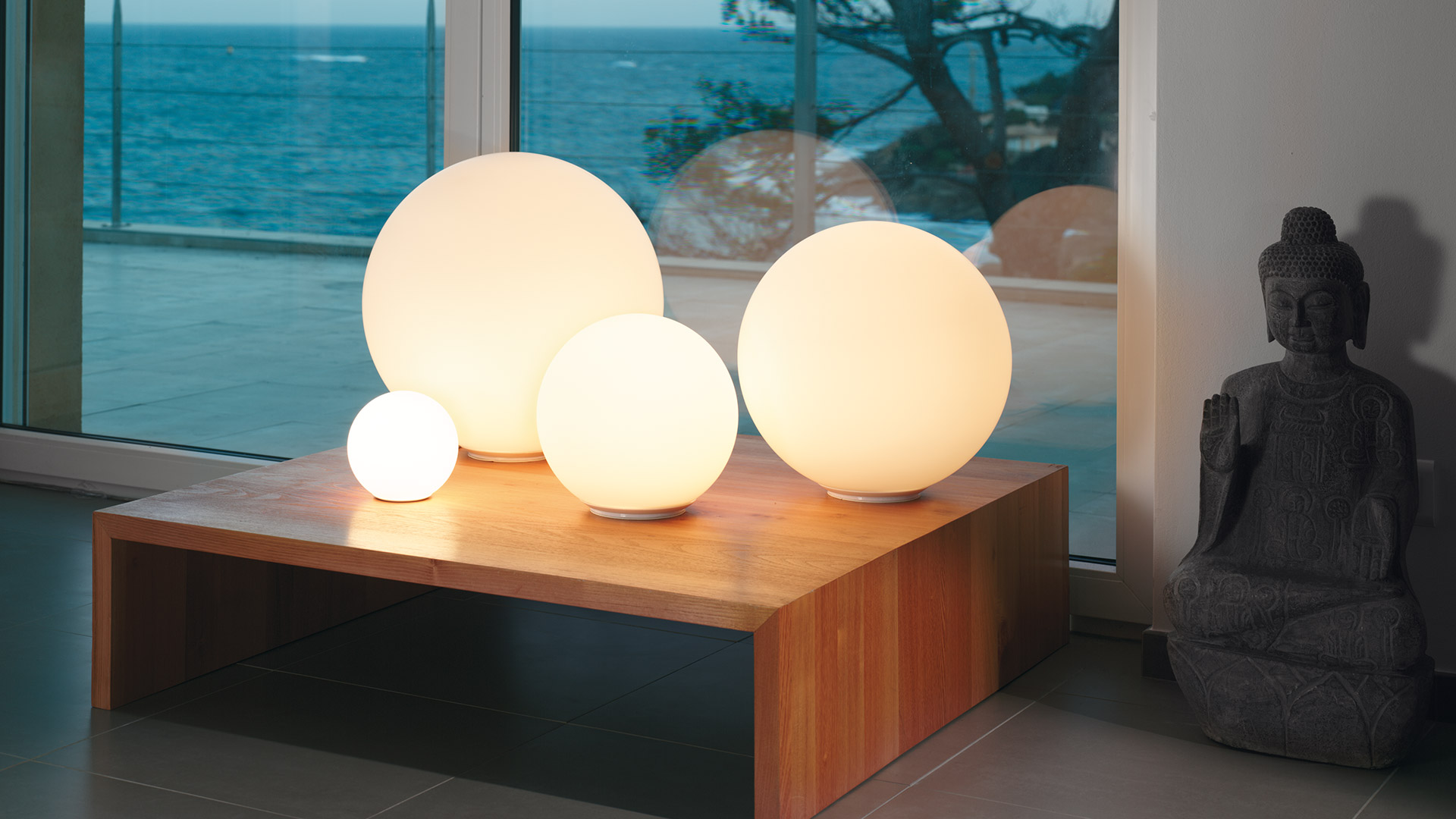 Quattro lampade da tavolo Artemide Dioscuri in vetro opalino bianco a sfera di diverse dimensioni disposte su una superficie bianca davanti a una parete giallo caldo