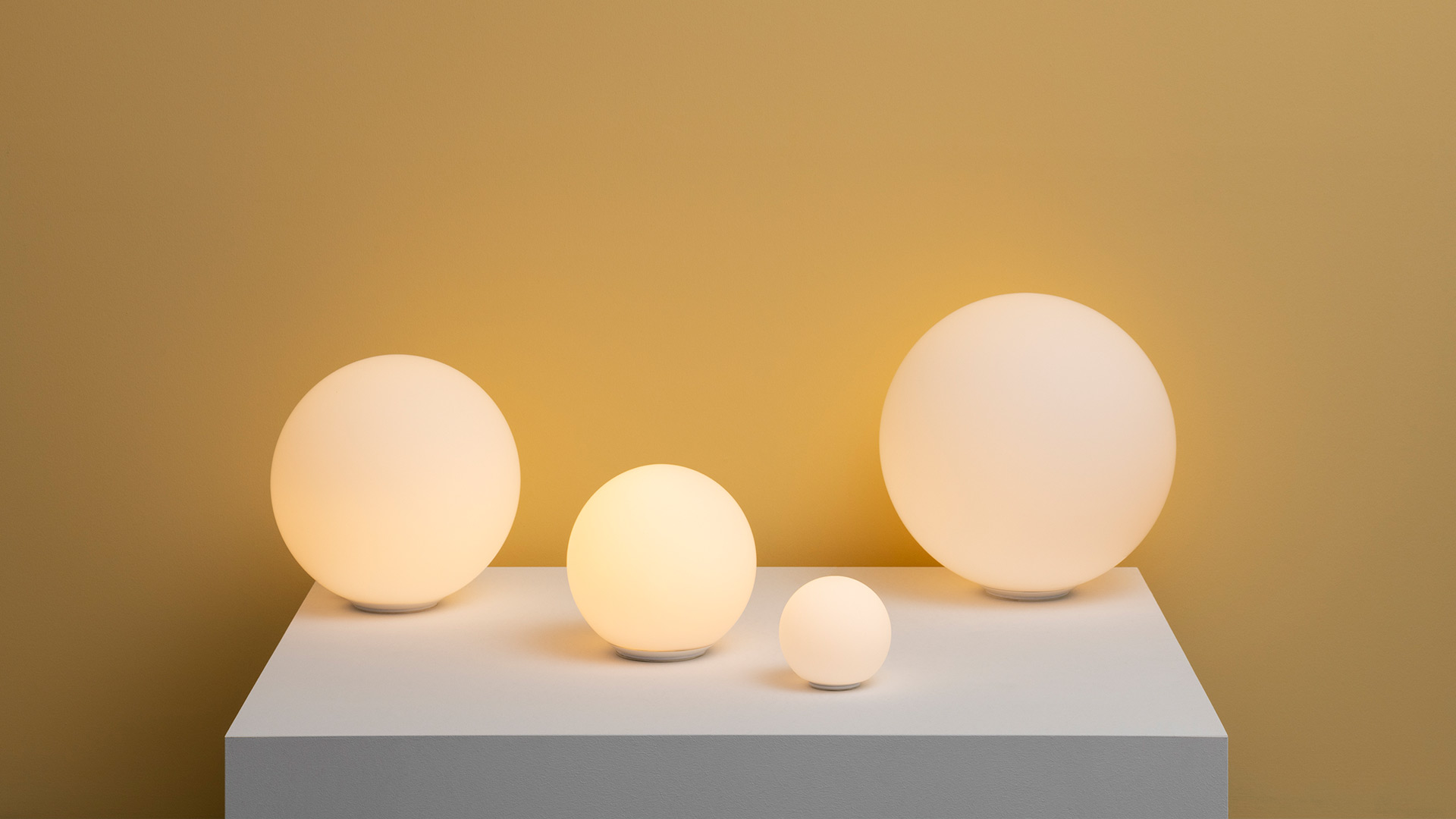 Quattro lampade da tavolo Artemide Dioscuri in diverse dimensioni, sfere di vetro opalino bianco che emanano luce calda su una piattaforma in legno vicino a una finestra sul mare