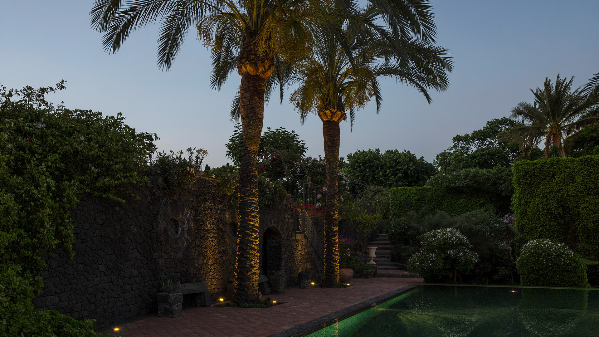 Giardino al crepuscolo con palme illuminate da faretti a terra Artemide Ego Spot, riflessi in una vasca accanto a un muro in pietra