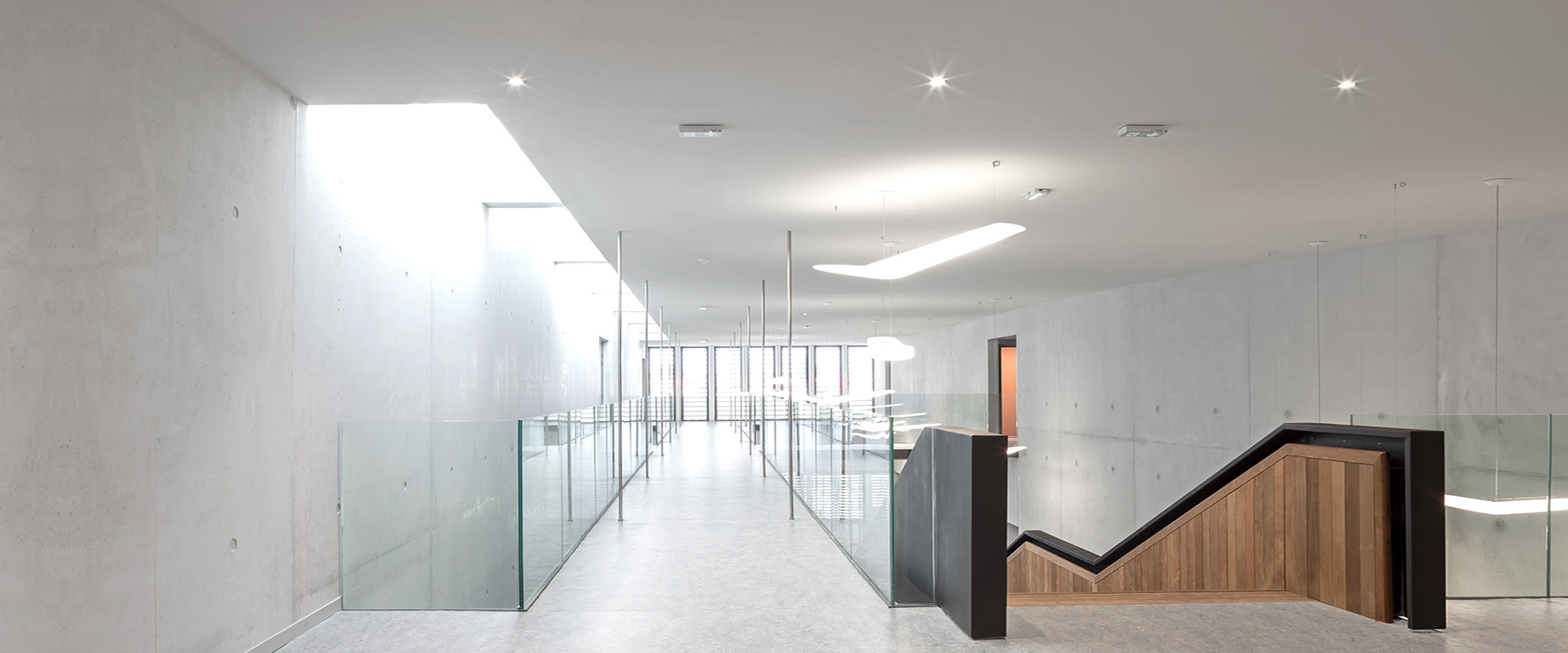 Corridoio moderno con faretti da incasso Artemide Everything 55 che illuminano pareti in cemento, ringhiere in vetro e una scala in legno.