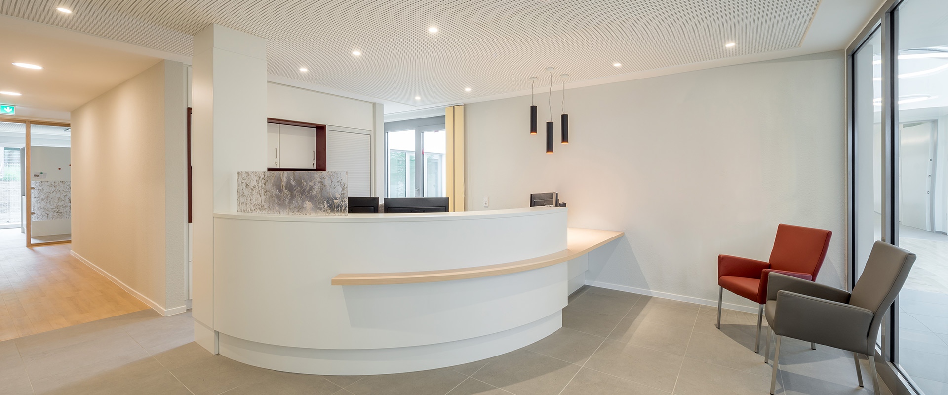 Area reception moderna con faretti da incasso Artemide Everything 80 Trimless e lampade a sospensione cilindriche scure sopra una scrivania bianca curva.
