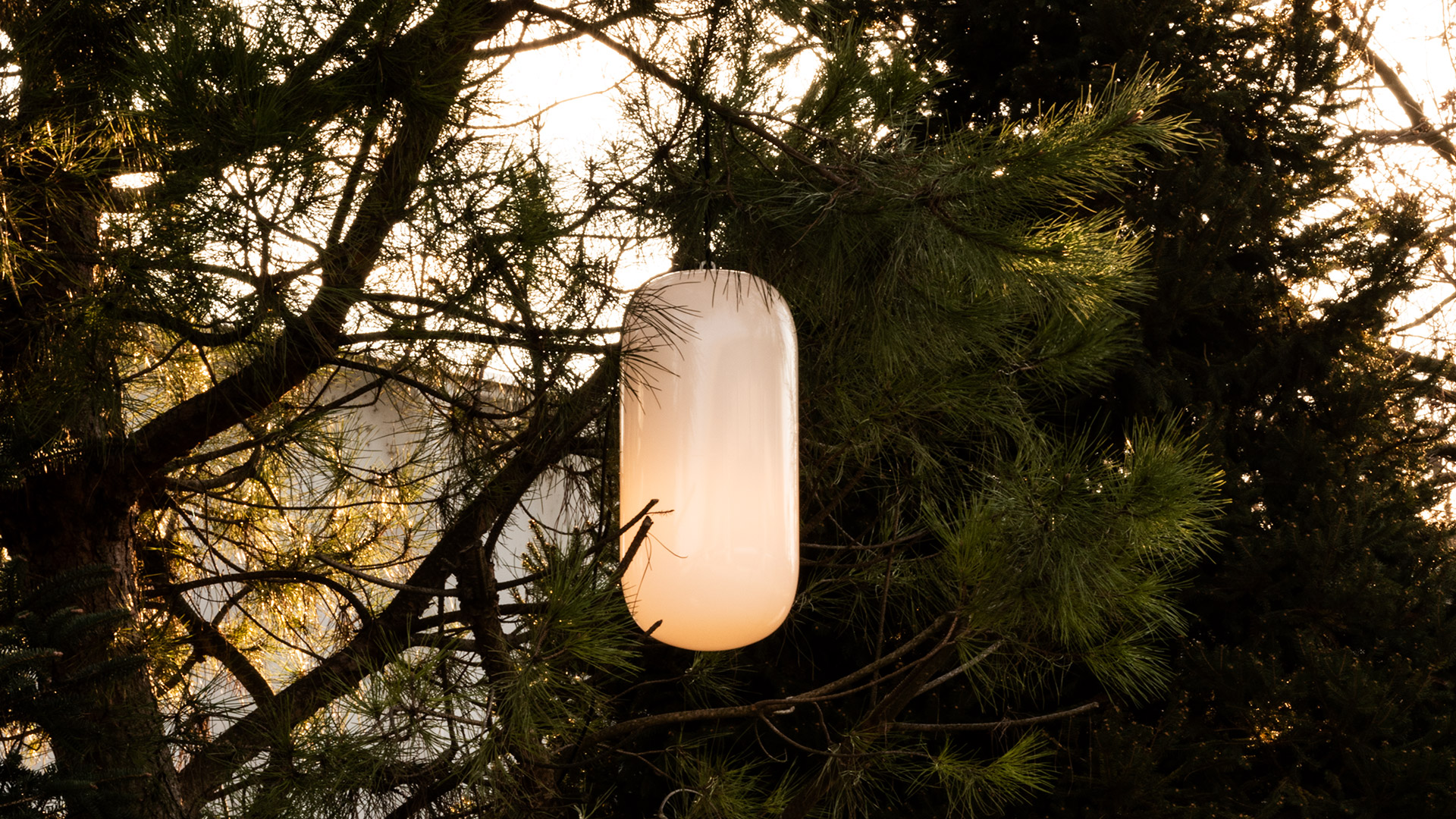 Lampada da terra Artemide Gople Outdoor con diffusore ovale bianco luminoso su palo in legno, posizionata accanto a un albero di notte vicino a un'amaca