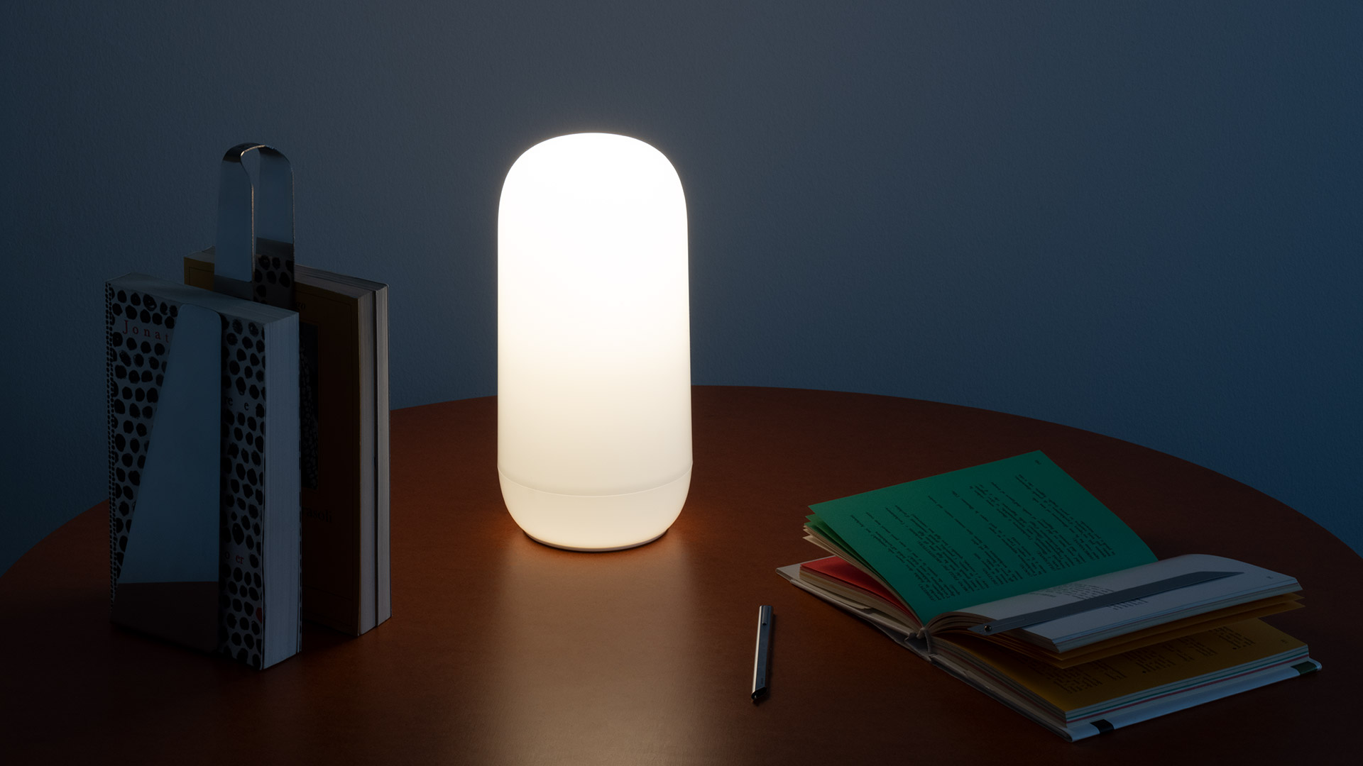 Lampada Artemide Gople Portable accesa su un tavolo rotondo in legno, accanto a libri e un taccuino, che diffonde luce calda in una stanza soffusa