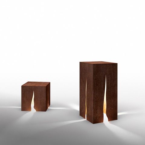 Due lampade da terra Artemide Granito in acciaio Corten, forme cubica e rettangolare alta, con luce calda che filtra attraverso fessure naturali