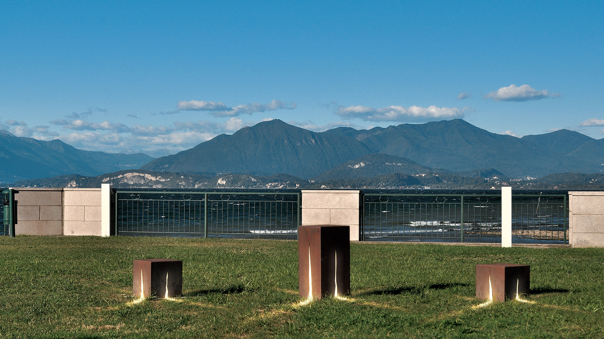Installazione outdoor Artemide Granito: elementi cubici scuri emettono fasci di luce rossa intensa su un pavimento riflettente, creando un motivo geometrico a stella.