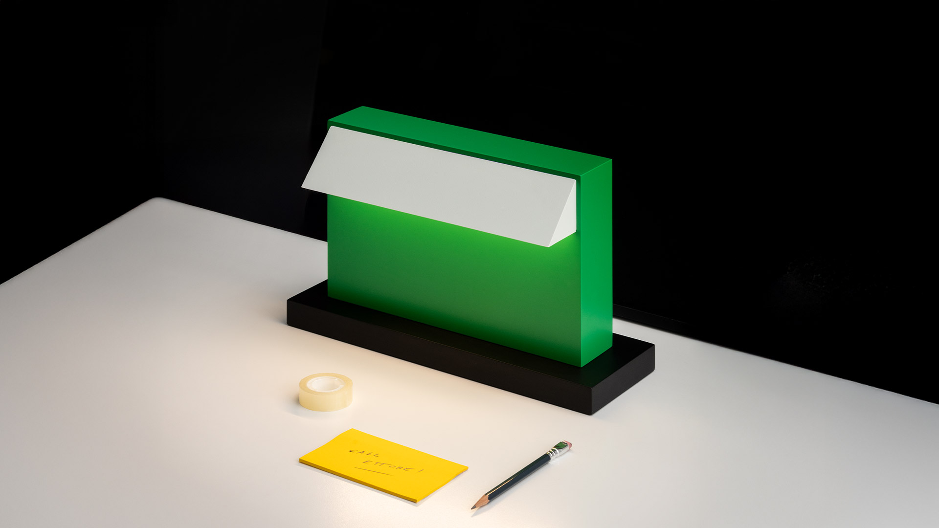 Lampada da tavolo Artemide Hera: corpo rettangolare verde compatto con riflettore bianco inclinato su base nera, mostrata accanto a una mela rossa per scala