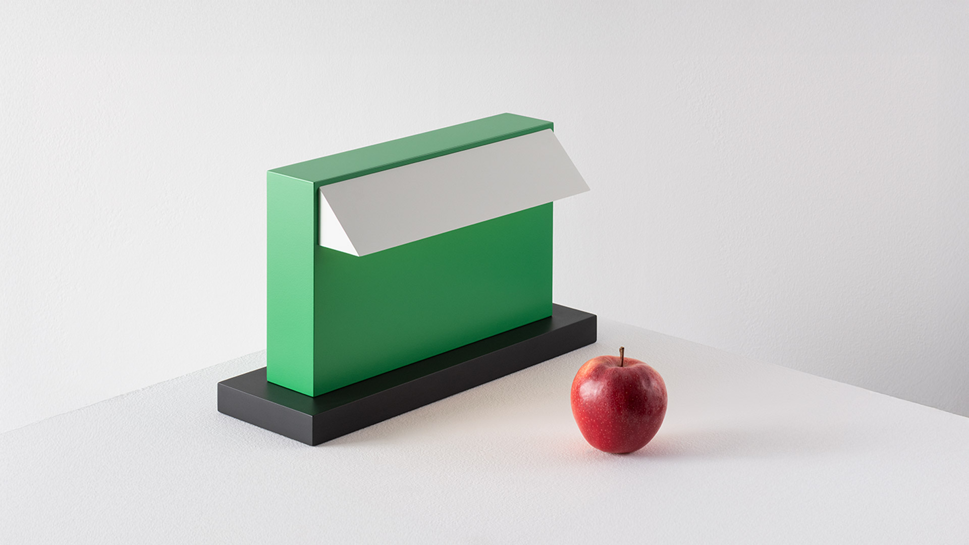 Lampada da tavolo Artemide Hera in verde brillante con diffusore bianco su base nera, illumina una scrivania con blocco note e penna
