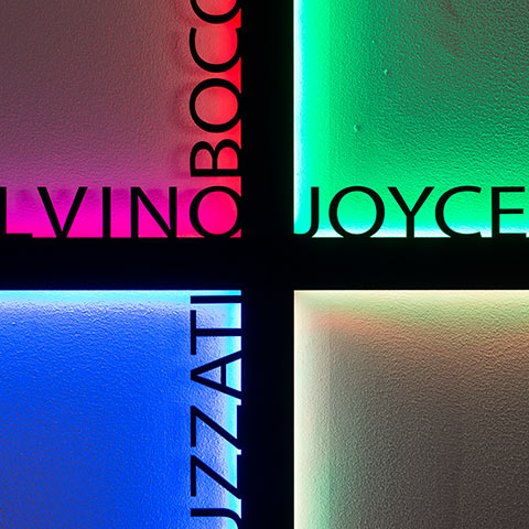 Four illuminated frosted glass panels with vibrant RGB colors—pink, green, blue, white—divided by dark cross frame, displaying literary names