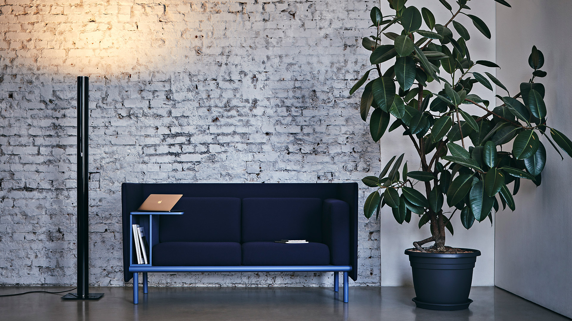Lampada da terra Artemide Ilio nera accanto a un divano blu navy con mensola in legno, su sfondo di muro di mattoni bianchi con pianta in vaso