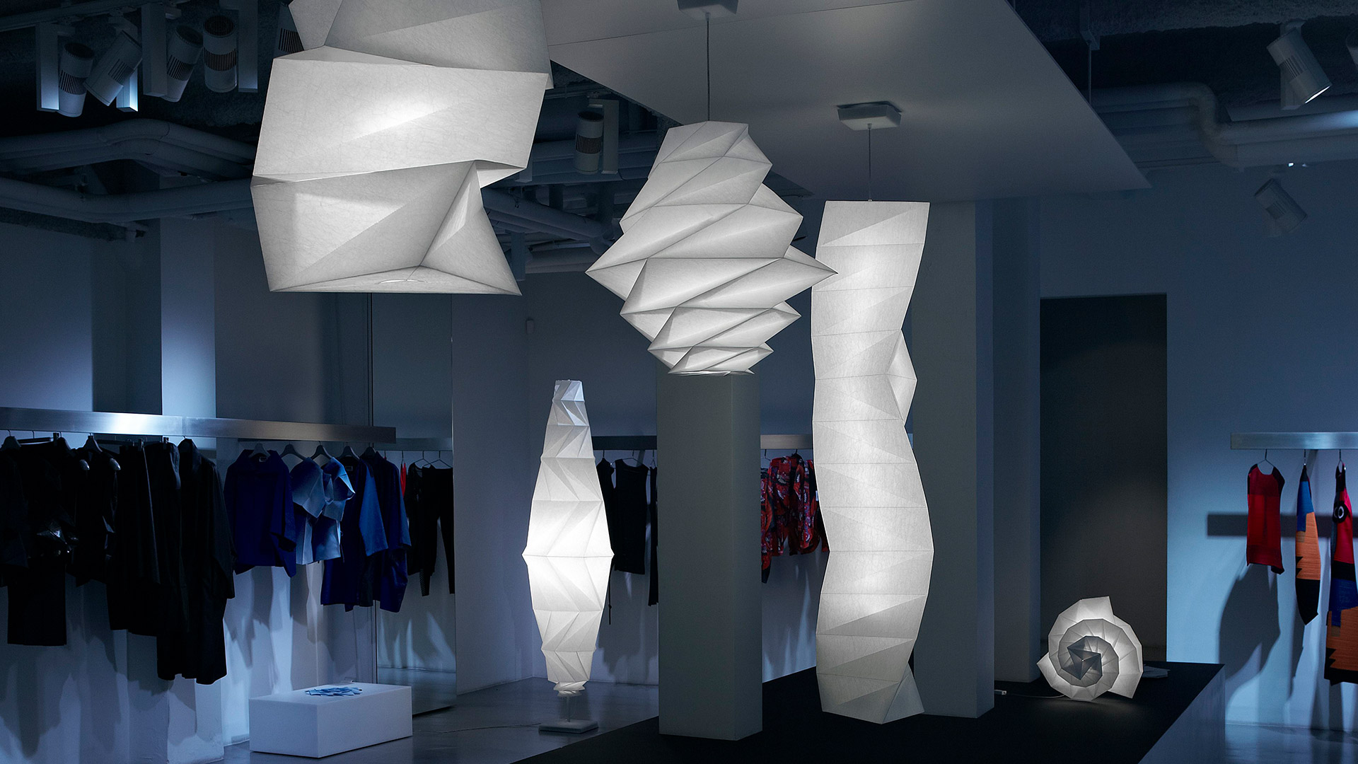 Tre lampade Artemide In-Ei Issey Miyake: due sospensioni a pieghe origami e un elemento a spirale a terra, luminosi bianchi su sfondo nero