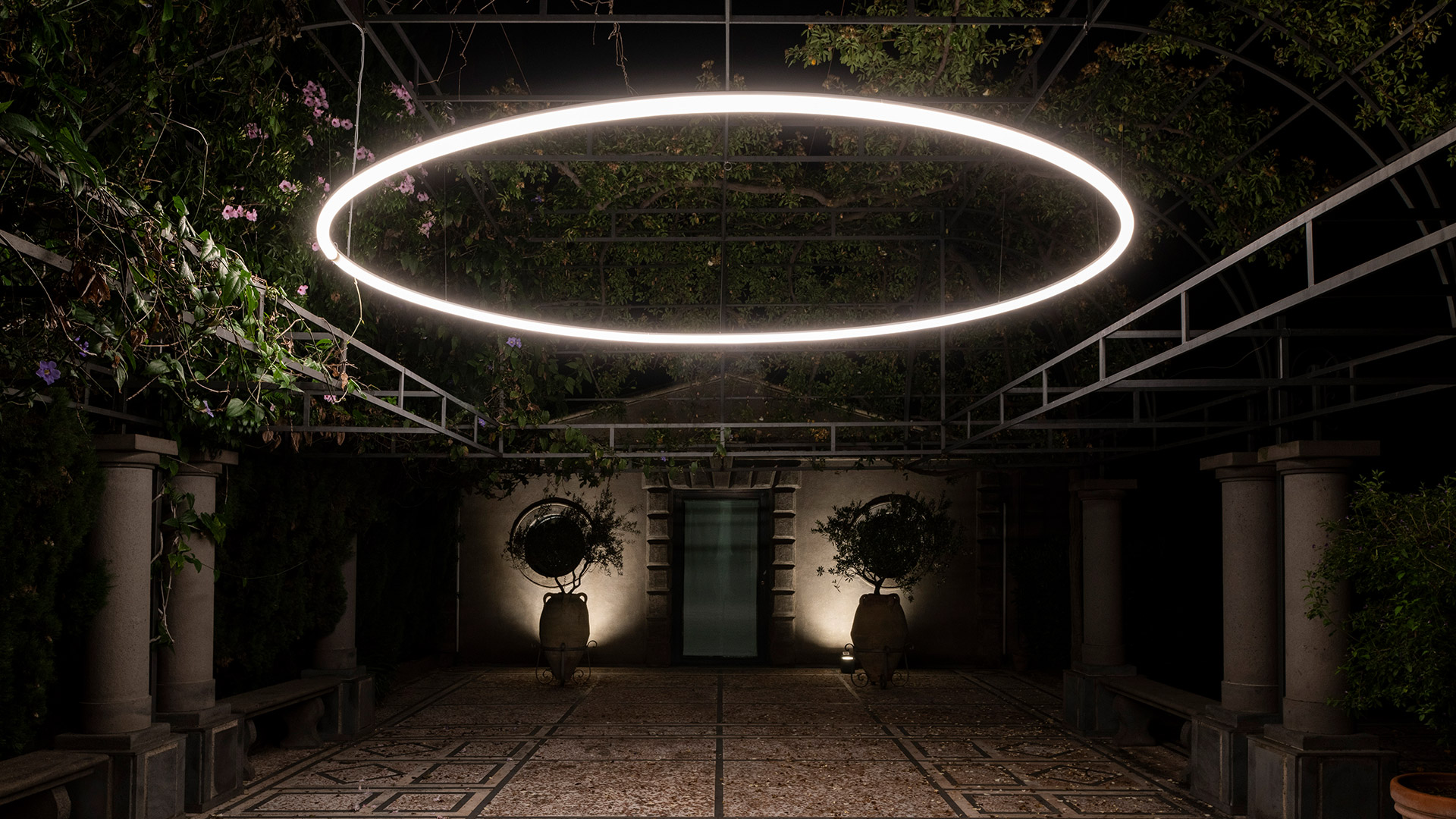 Lampada a sospensione Artemide La Linea con forma a nastro luminoso sinuoso, sospesa tra i rami di un glicine rigoglioso in una pergola esterna
