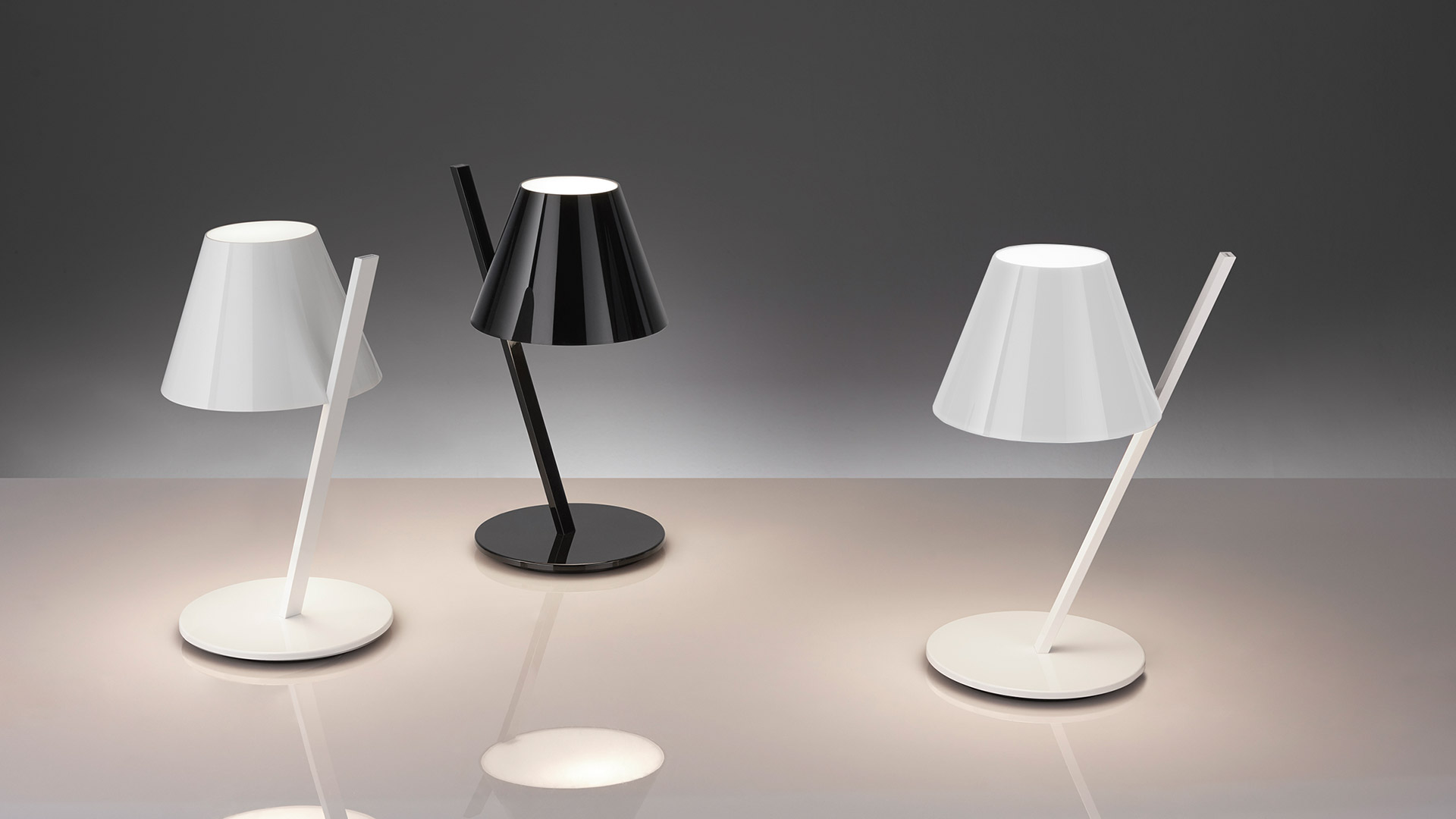 Lampada da tavolo Artemide La Petite bianca con paralume conico su stelo inclinato, illumina calorosamente una scrivania in legno con libri e pianta