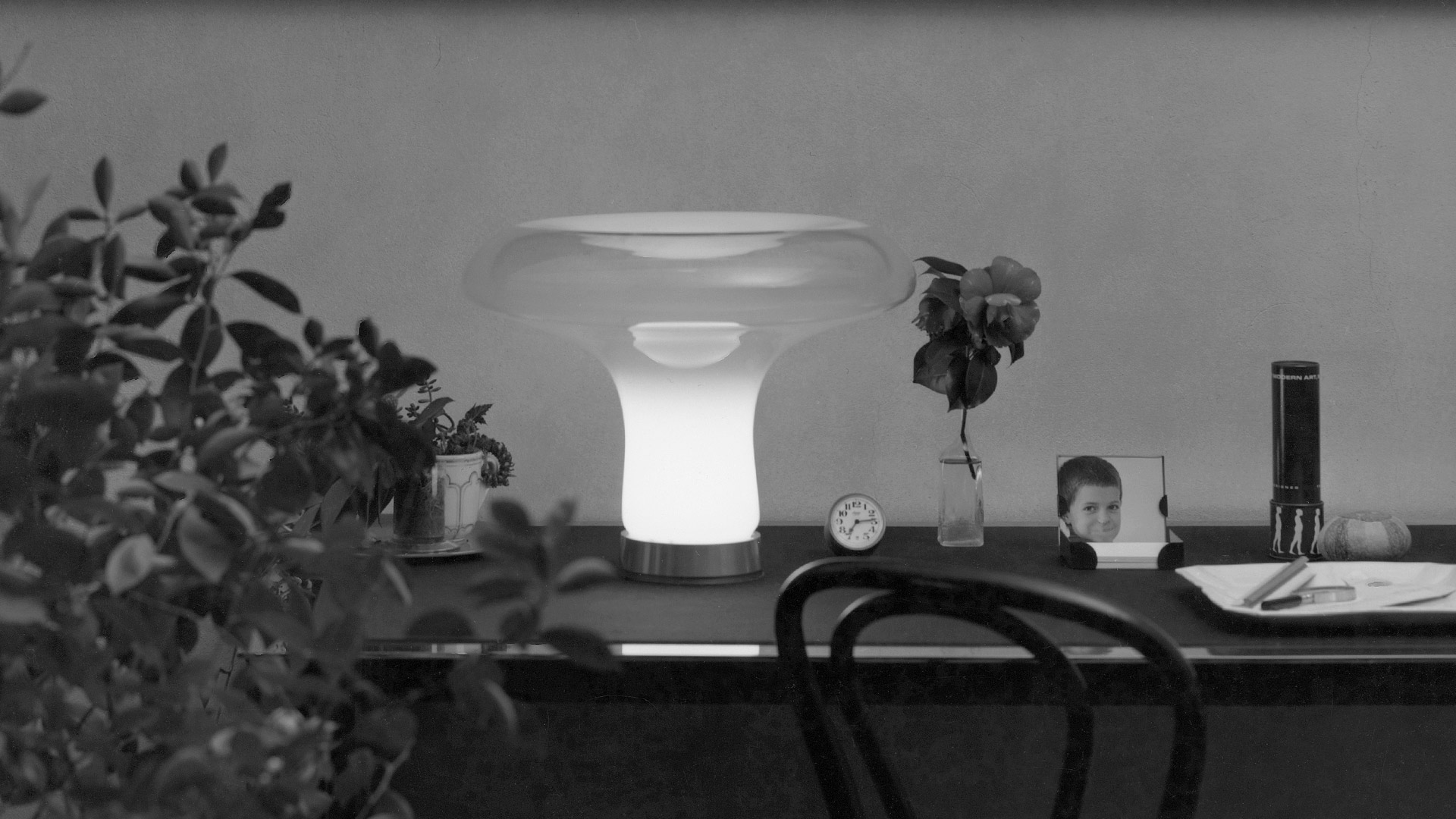 Lampada da tavolo Artemide Lesbo con diffusore in vetro satinato a forma di fungo e base cromata, illuminata calorosamente su un piedistallo bianco