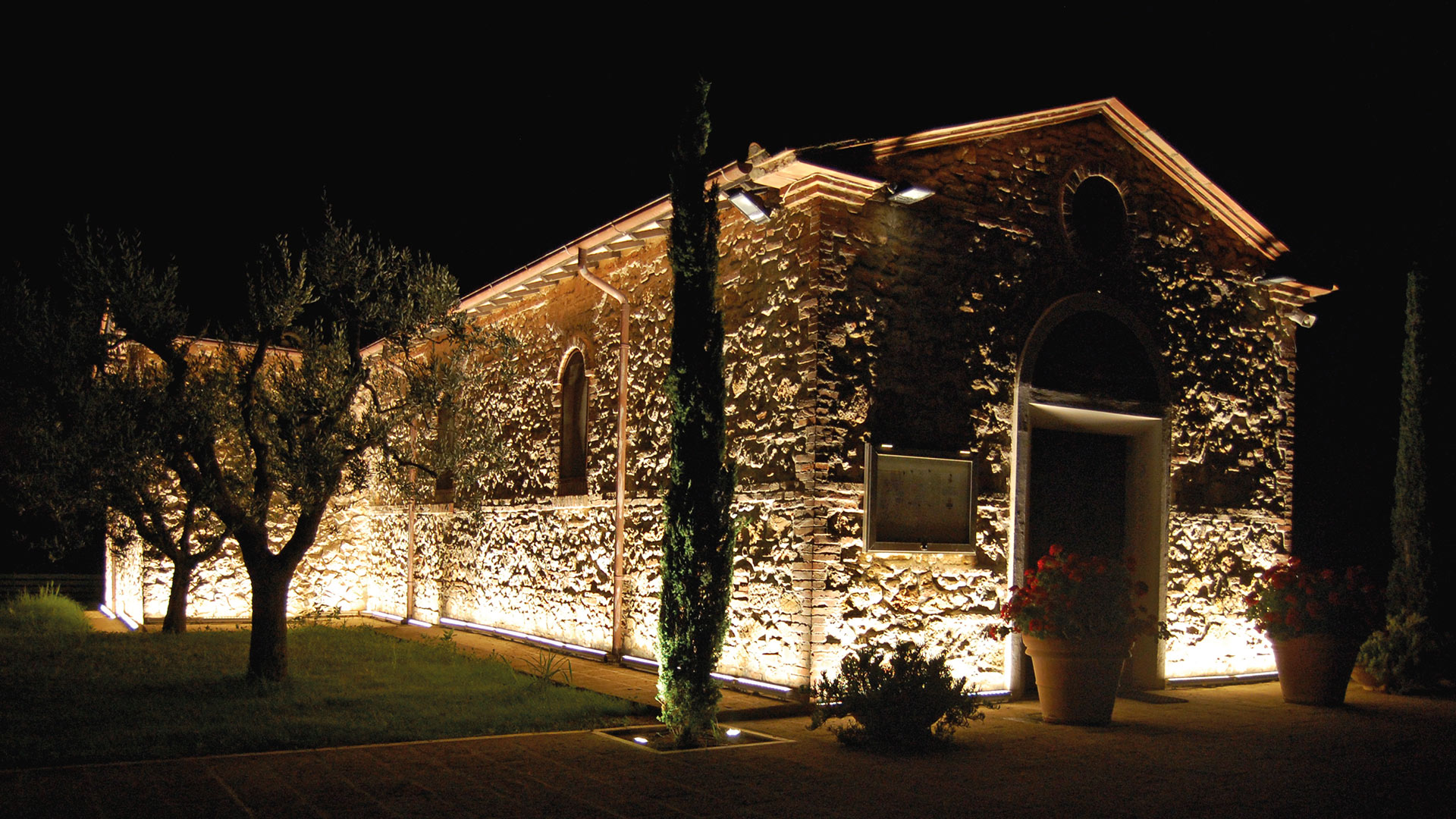 Cappella storica in pietra illuminata di notte da apparecchi Artemide LineaLed Wallwasher, che proiettano luce dorata calda su pareti e giardino