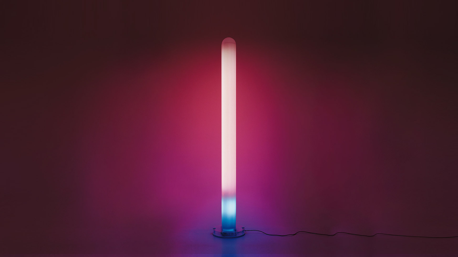 Lampada da terra Artemide Metacolor: alto tubo cilindrico che emette luce sfumata dal basso ciano al rosa-magenta in alto su sfondo scuro