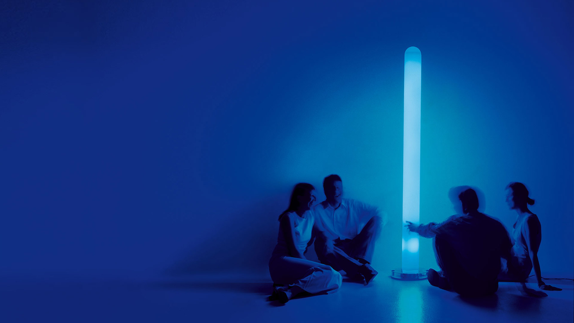 Quattro persone sedute attorno alla lampada da terra cilindrica Artemide Metacolor che emette luce blu in una stanza illuminata di blu