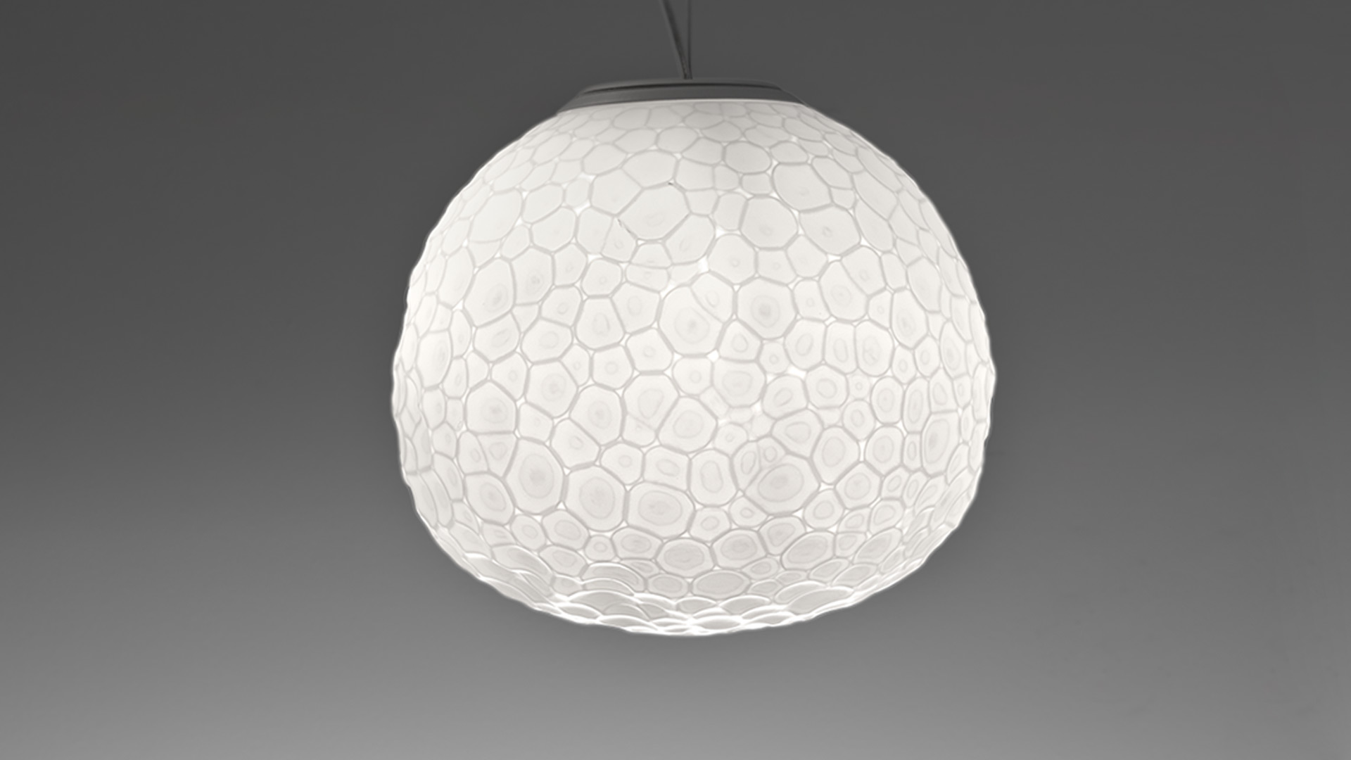 Lampada a sospensione Artemide Meteorite: diffusore sferico bianco con texture a crateri, emette luce calda e diffusa su sfondo scuro