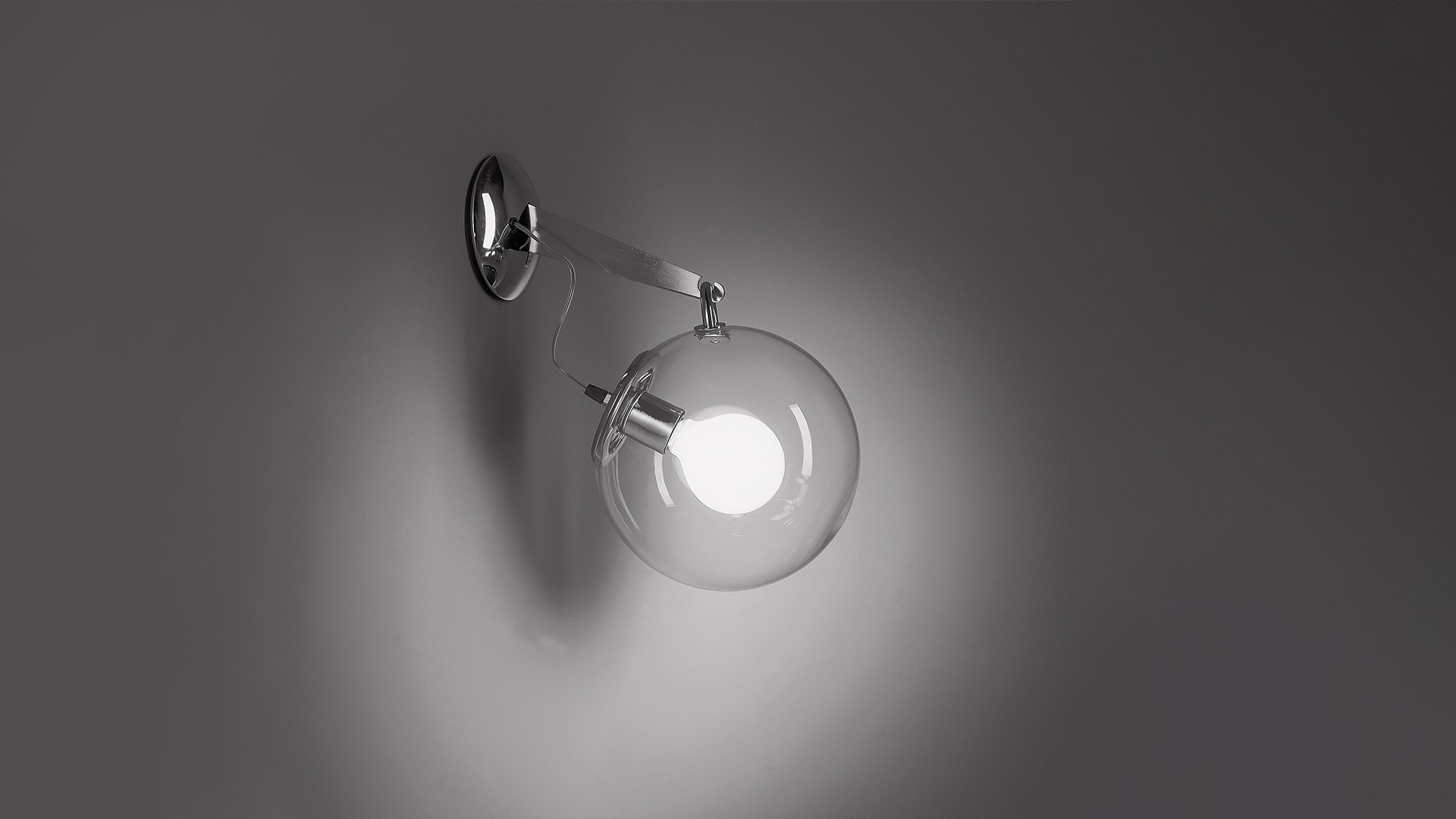Lampada da parete Artemide Miconos con braccio in ottone spazzolato e diffusore sferico in vetro trasparente, montata su parete bianca