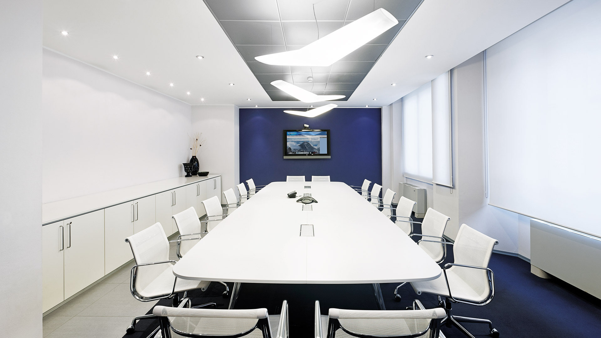 Sala conferenze moderna con lampade a sospensione Artemide Mouette sopra un lungo tavolo bianco circondato da sedie bianche, parete blu.