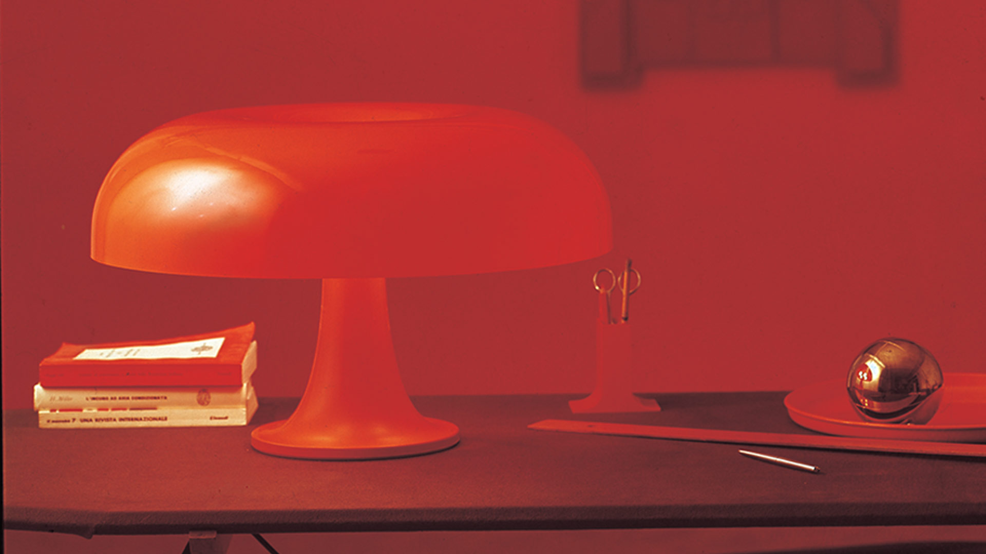 Lampada da tavolo Artemide Nesso in rosso, con l'iconico paralume a fungo su base a tromba, che emana una luce calda su un piedistallo bianco