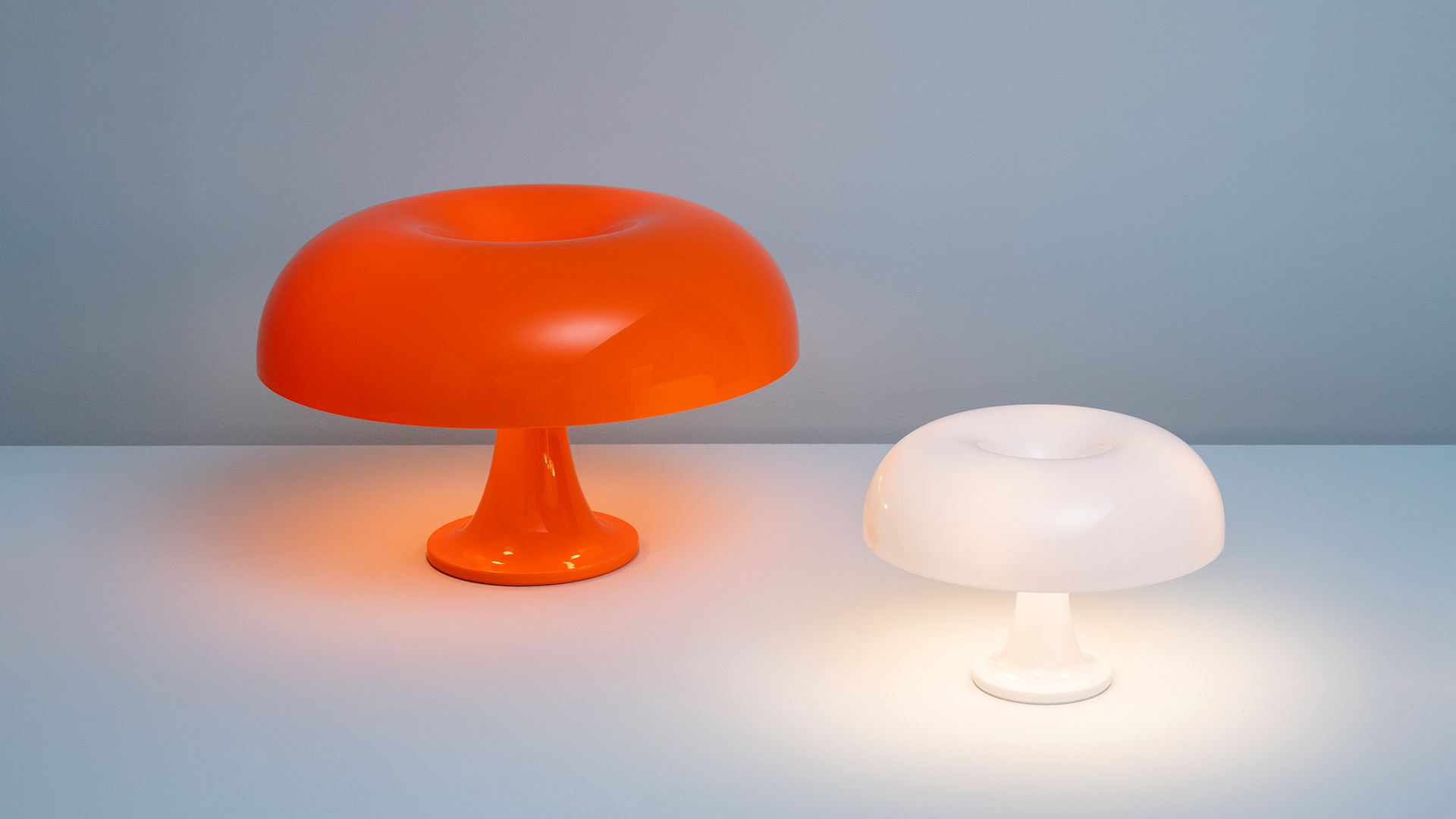 Due lampade da tavolo Artemide Nesso: grande lampada a fungo arancione a sinistra, versione bianca illuminata più piccola a destra