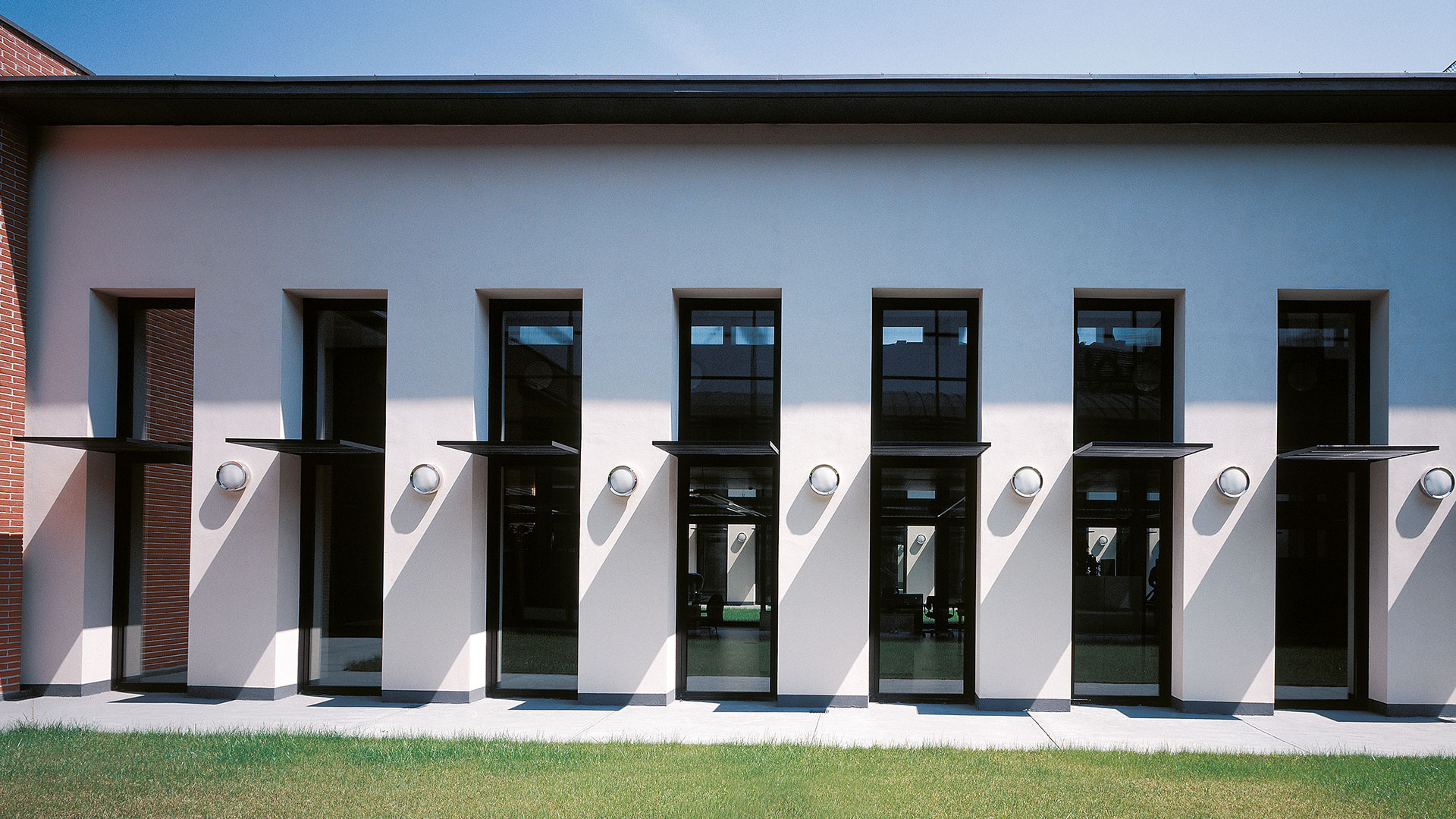 Facciata di edificio moderno con alte finestre a telaio scuro e applique da parete Artemide Niki montate tra ogni campata