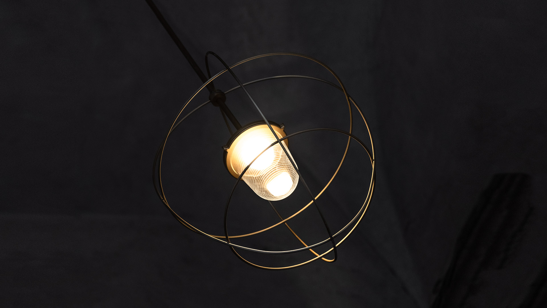 Lampada a sospensione Nottola con anelli orbitali in filo metallico dorato che incorniciano una lampadina opaca accesa, sospesa da cavo nero su sfondo scuro