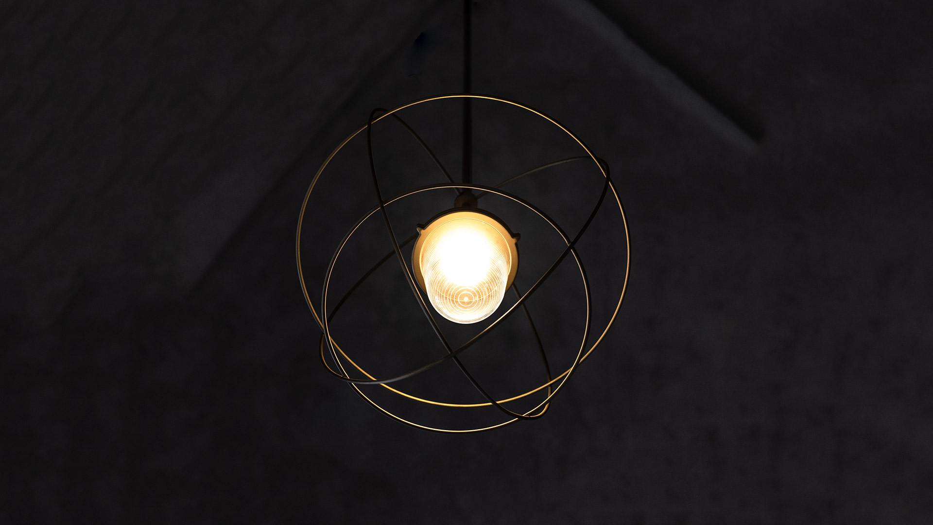 Lampada a sospensione Artemide Nottola: bulbo a vista racchiuso in anelli metallici dorati incrociati, sospesa su soffitto scuro