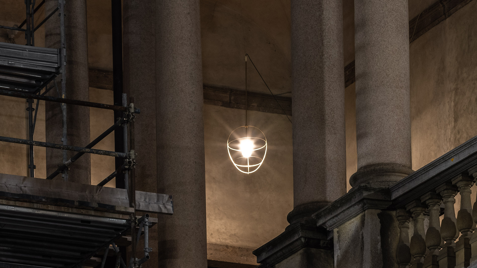 Lampada a sospensione Artemide Nottola con struttura a gabbia bianca e bulbo a vista, appesa tra colonne in pietra classiche in un edificio storico