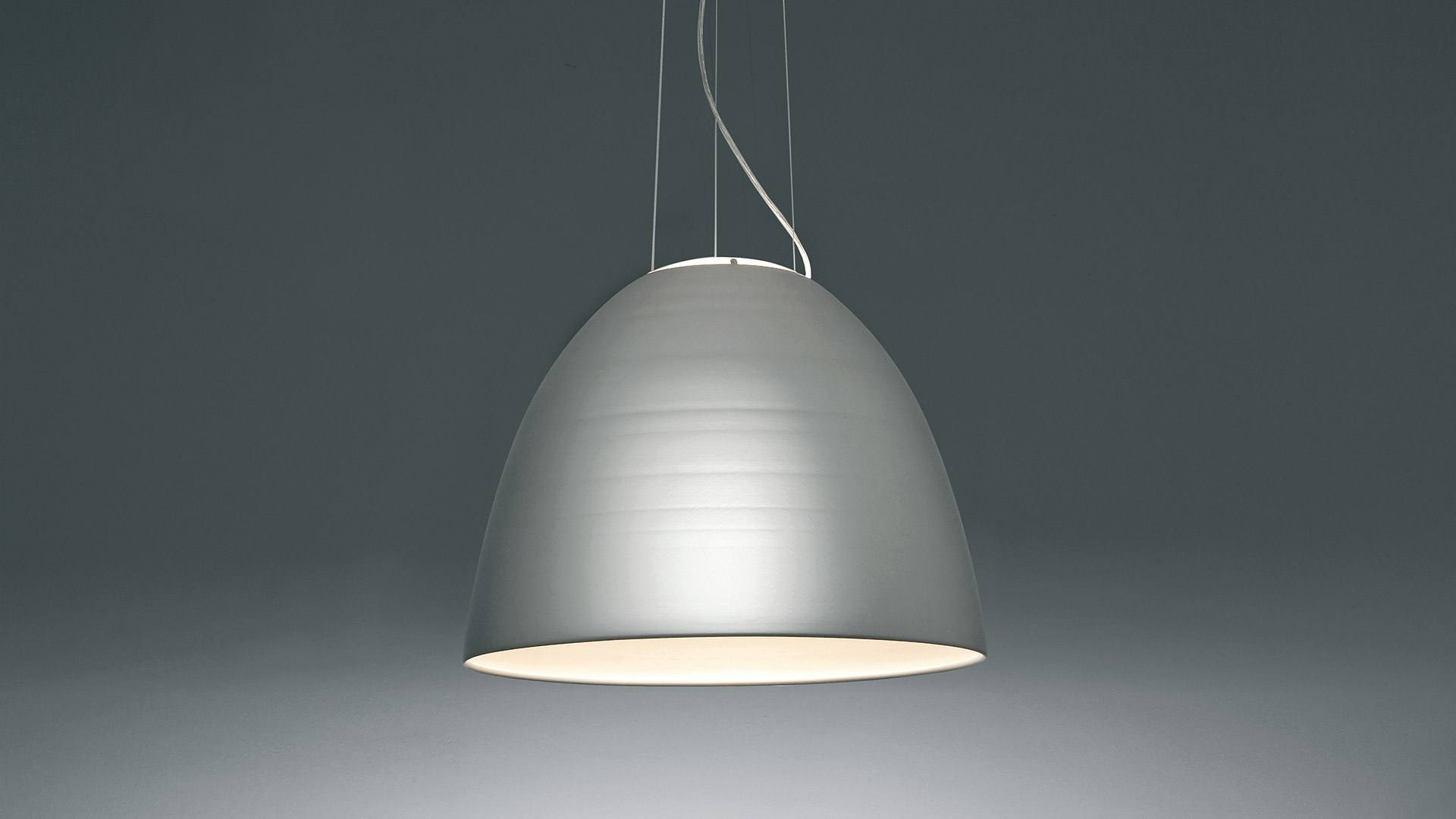 Lampada a sospensione Artemide Nur con paralume a cupola antracite scuro, interno bianco, sospesa da sottili cavi su sfondo grigio