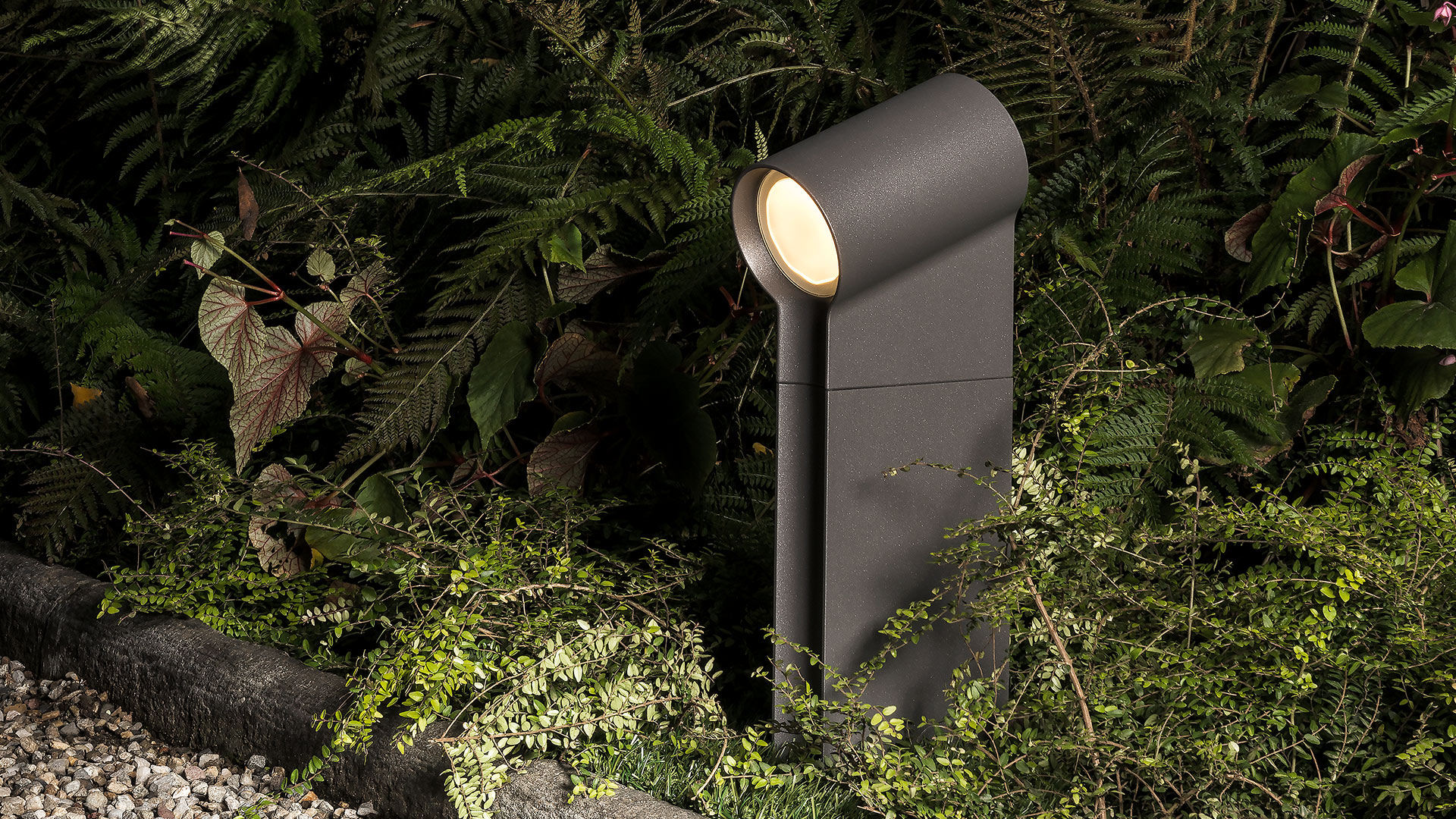 Lampada da esterno Artemide Oblique color grigio scuro, con testa LED inclinata che illumina un rigoglioso giardino di notte