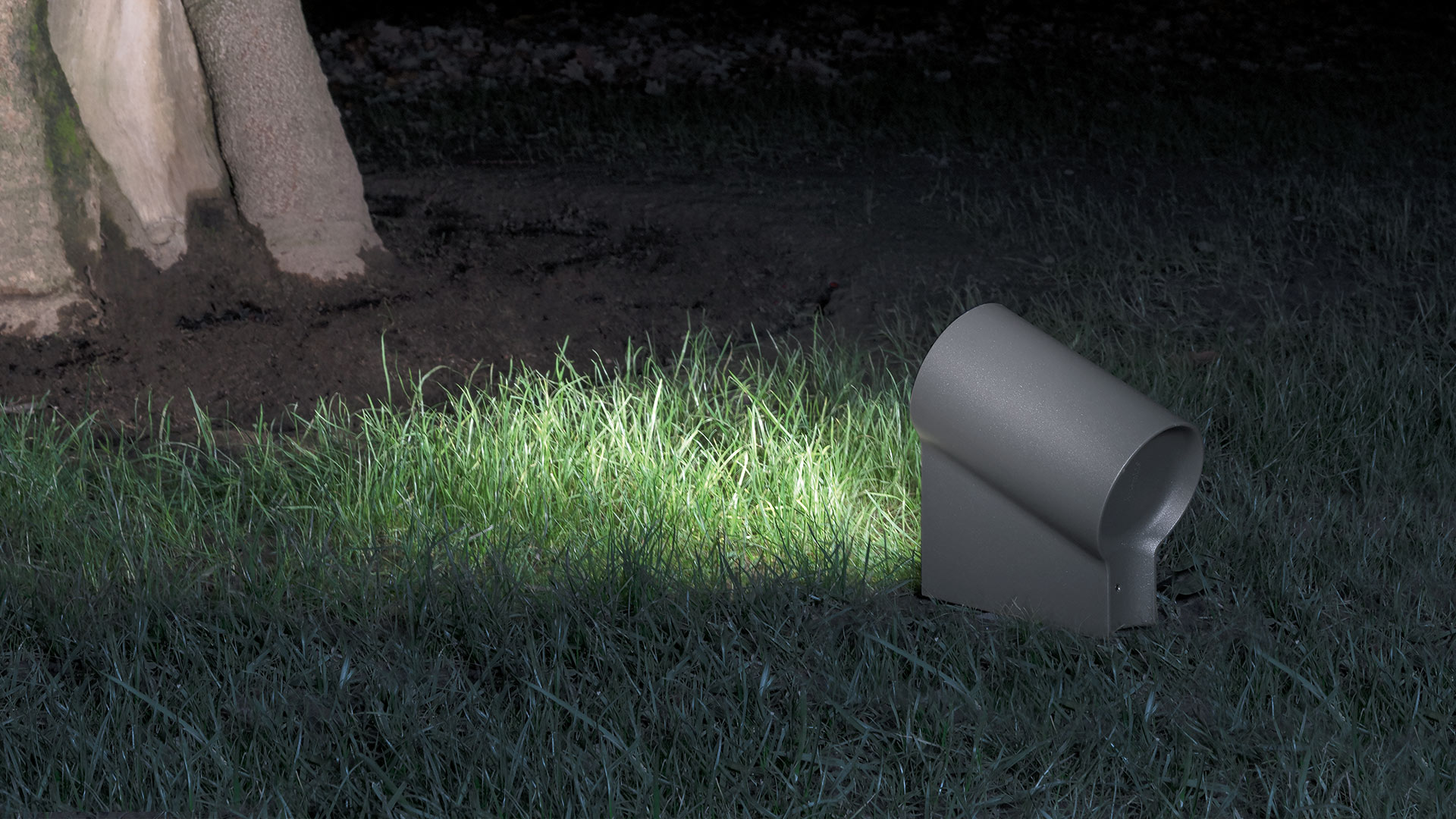 Faretto da esterno Artemide Oblique grigio scuro, installato a terra su prato, con luce concentrata verso la base di un albero di notte