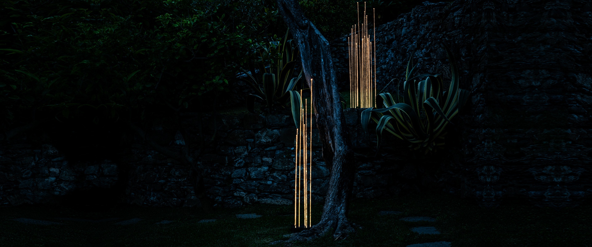 Lampada da terra Artemide Reeds Outdoor: fascio di sottili aste verticali bianche illuminate di notte su sfondo di fiori rossi