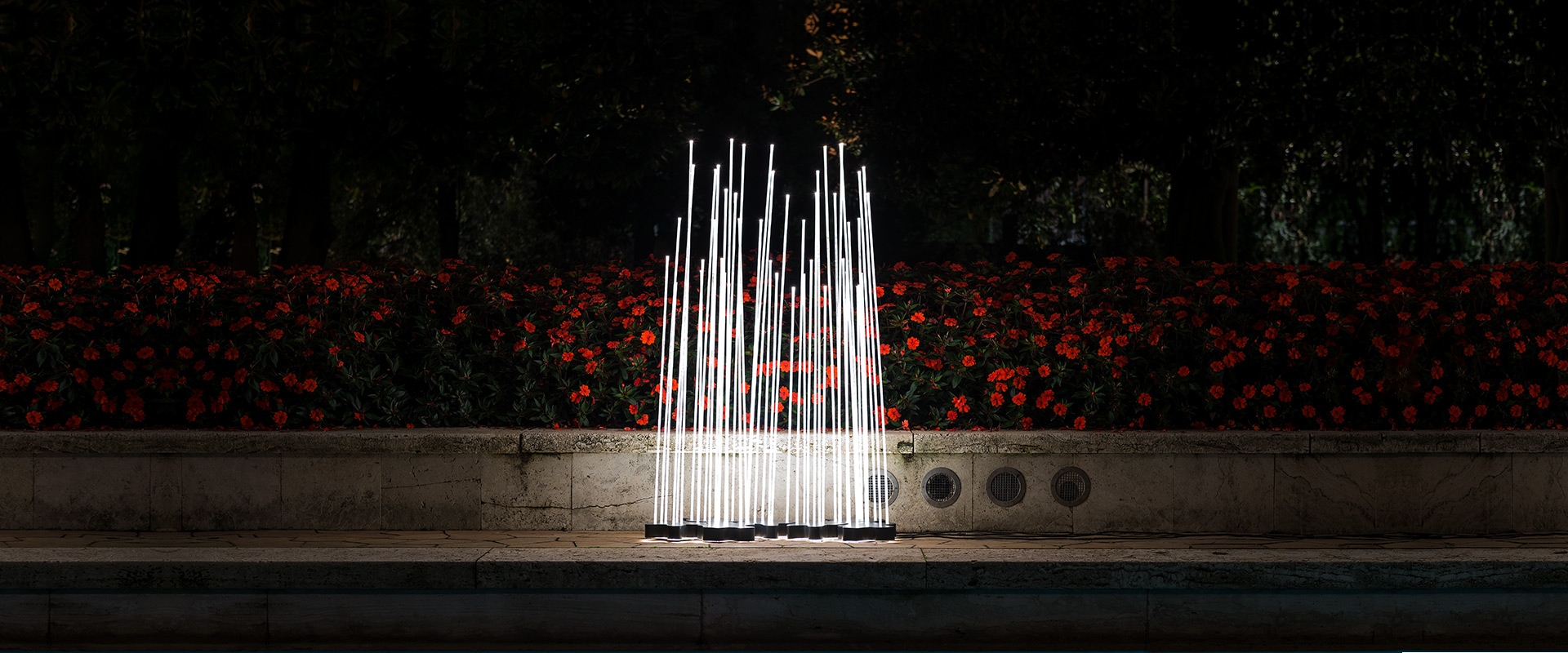 Lampada da terra Artemide Reeds Outdoor: più canne verticali dorate illuminate disposte come canne palustri, accanto a un albero in un giardino notturno