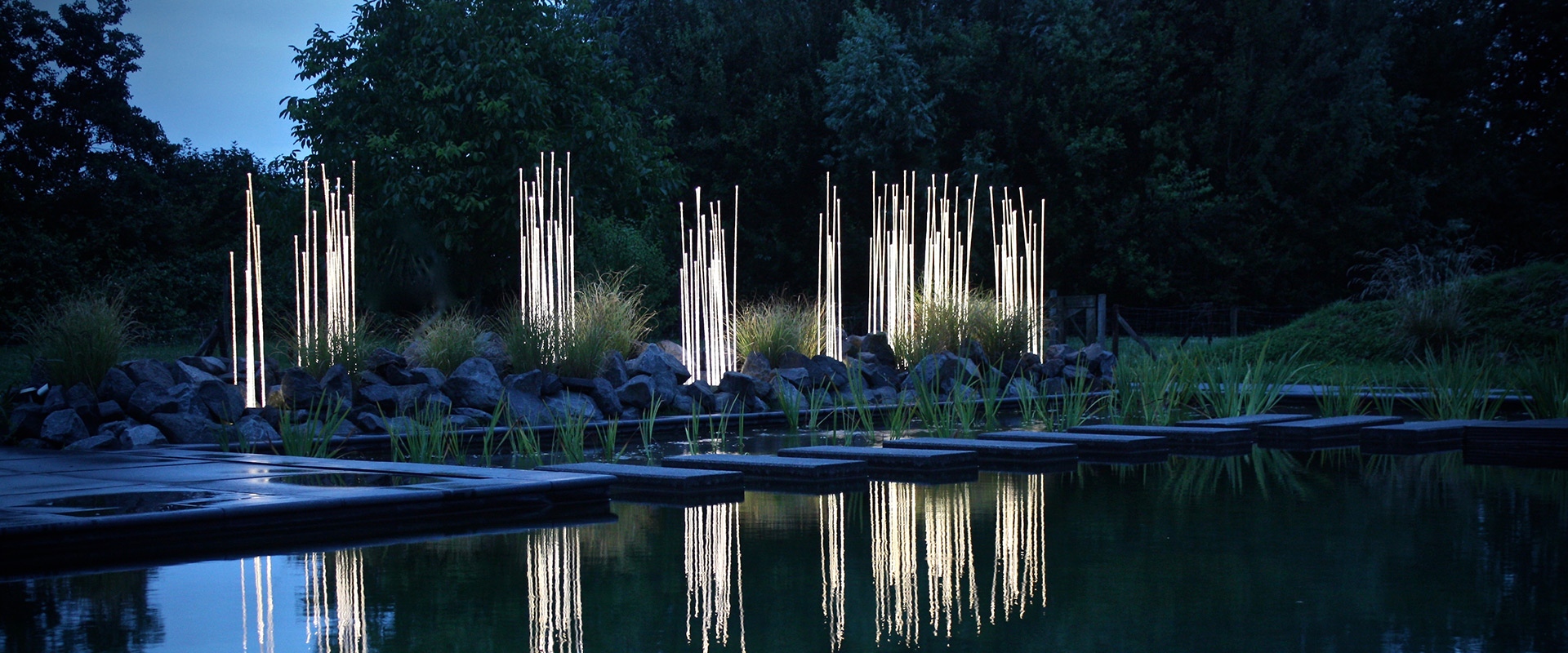 Lampade Artemide Reeds Outdoor: alte colonne luminose sottili riflesse in uno specchio d'acqua, circondate da rigogliosa vegetazione al crepuscolo
